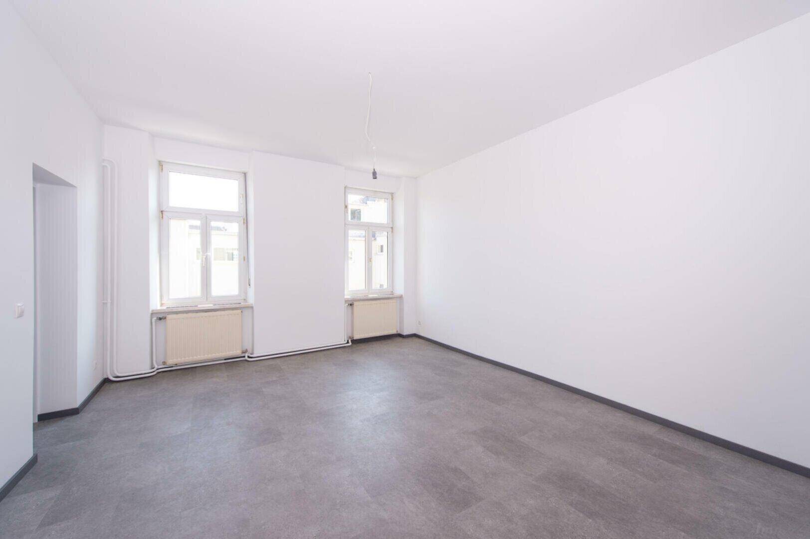 Geräumiger, heller Raum mit weißen Wänden, grauem Boden und zwei großen Fenstern.