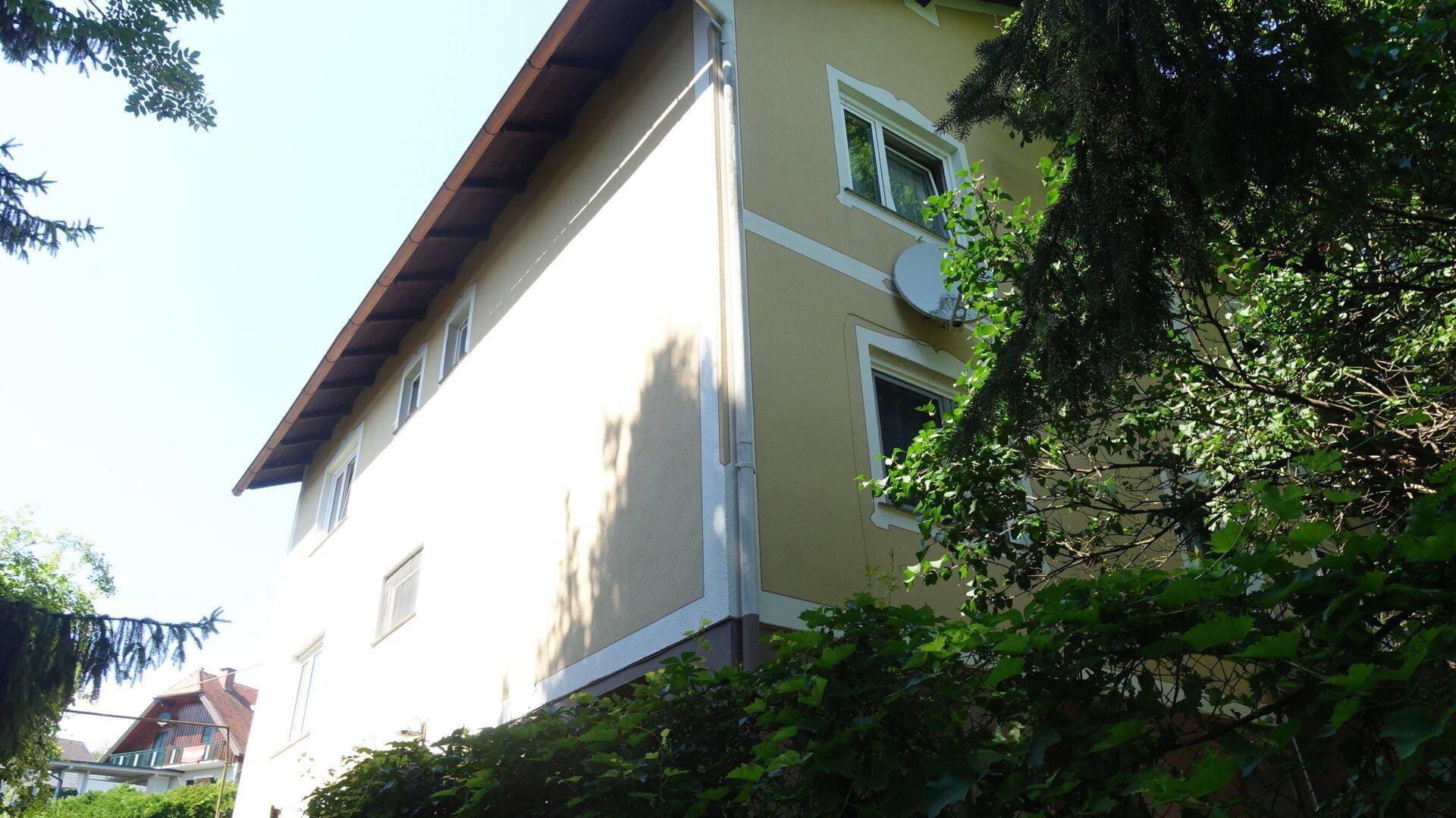 Seitenansicht des Hauses mit beiger Fassade und Fenstern, umgeben von Bäumen und Sträuchern.