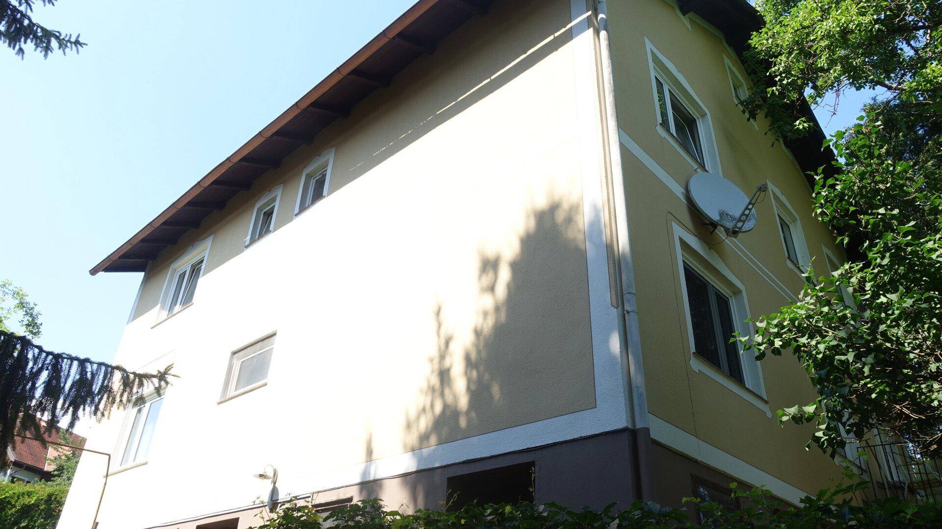 Seitenfassade des Hauses mit beigem Putz und Satellitenschüssel, umgeben von viel Grün.