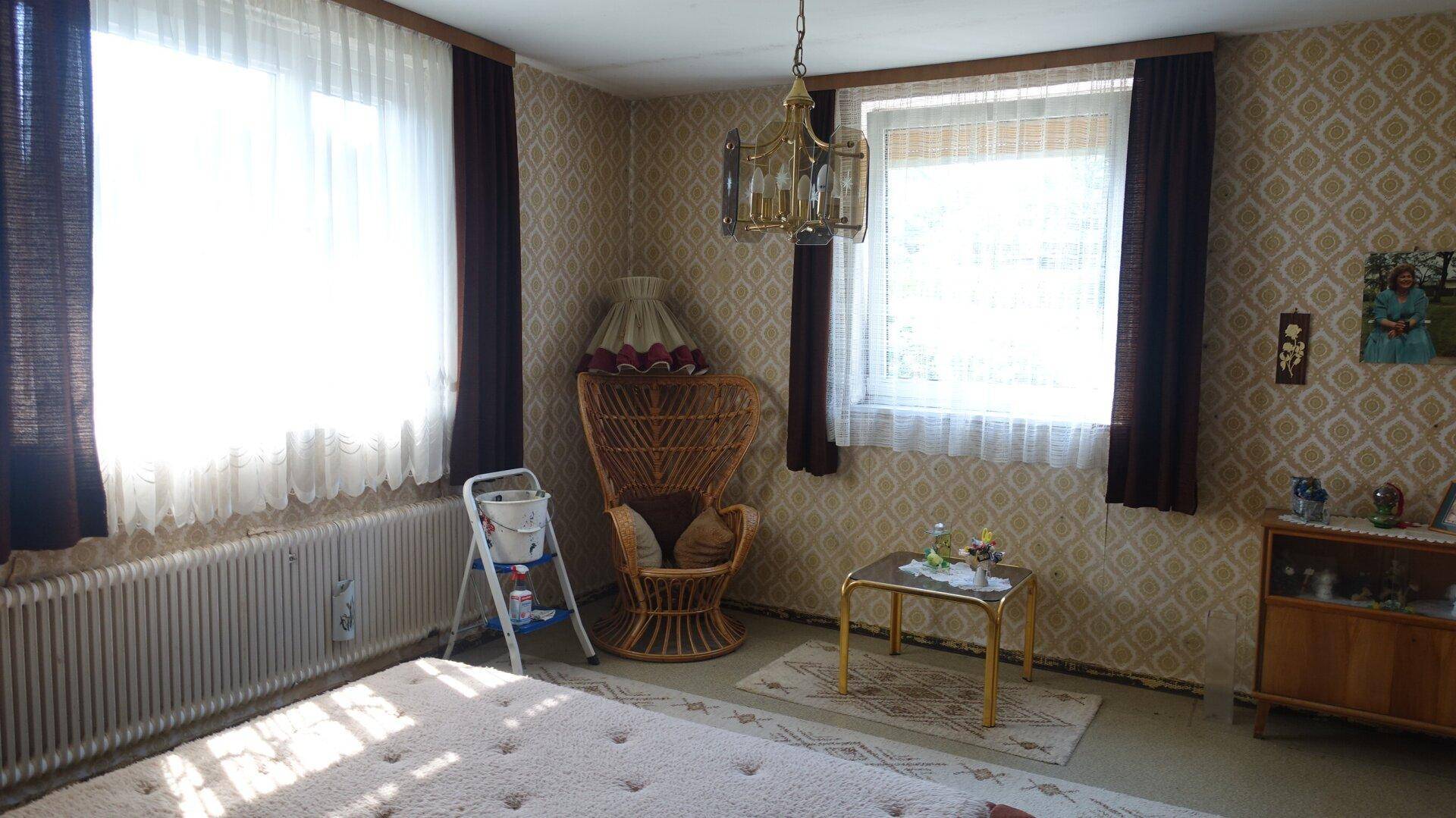 Schlafzimmer mit gemusterter Tapete, Rattan-Sessel und zwei Fenstern, traditionell eingerichtet.