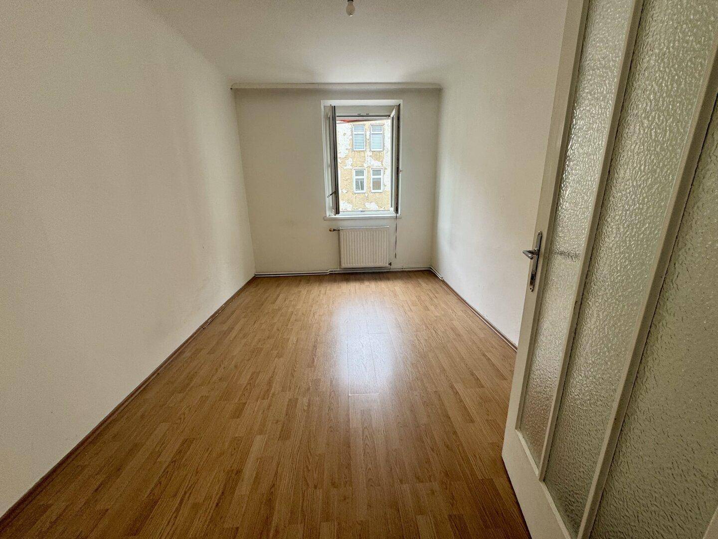 Leeres Zimmer mit Laminatboden und Fensterblick auf die umliegenden Gebäude.