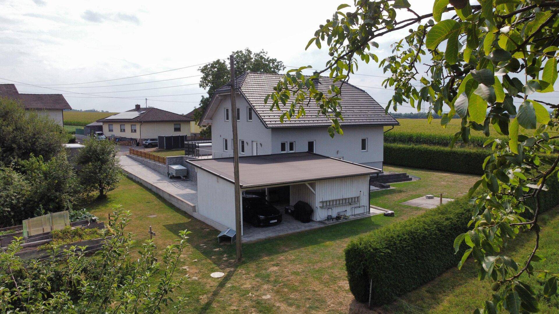 Seitenansicht des Hauses mit Carport und Zufahrt, umgeben von Bäumen und Grünflächen.
