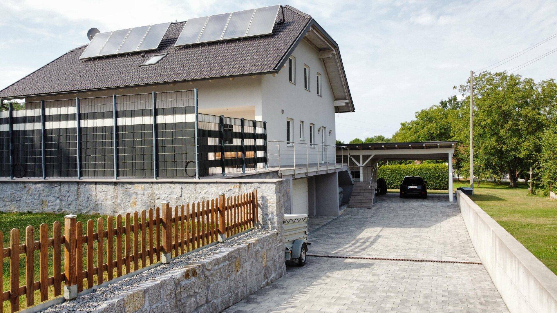 Seitenansicht des Hauses mit Garage, Carport und gepflasterter Zufahrt, umgeben von einem Holzzaun.