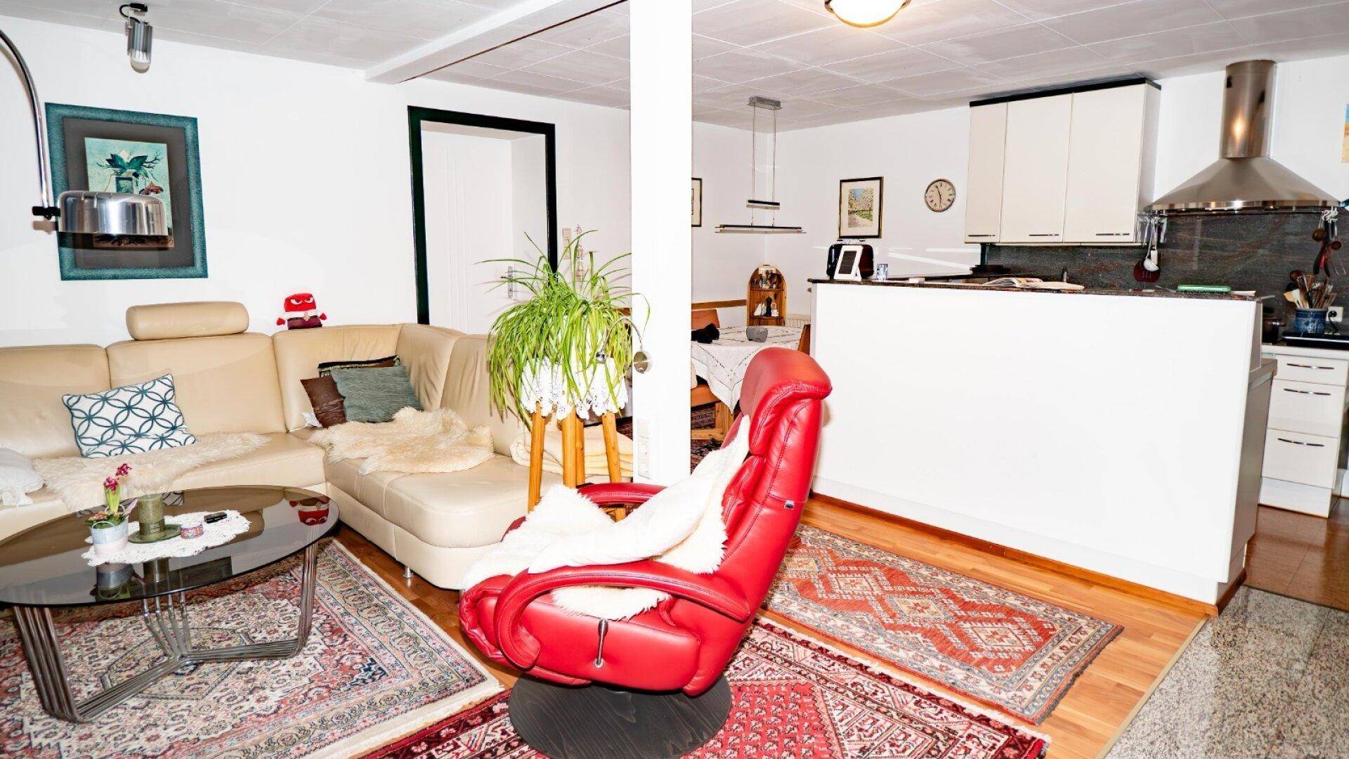 Gemütlicher Wohnbereich mit beiger Couch, rotem Sessel und offener Küche im Hintergrund.