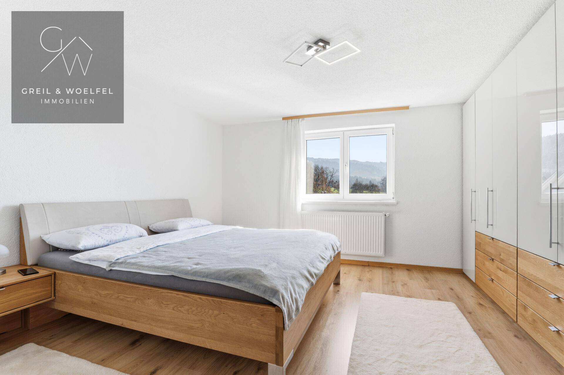 Helles Schlafzimmer mit Holzbett, weißem Kleiderschrank und einem Fenster mit Blick ins Grüne.