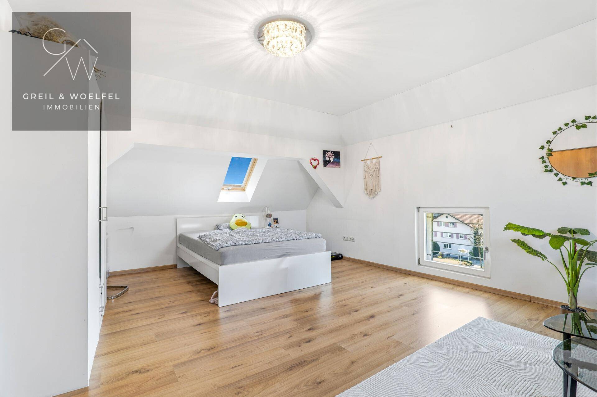 Helles Dachzimmer mit Bett, Holzboden, Dachfenster und dekorativer Deckenleuchte, bietet viel Tageslicht.