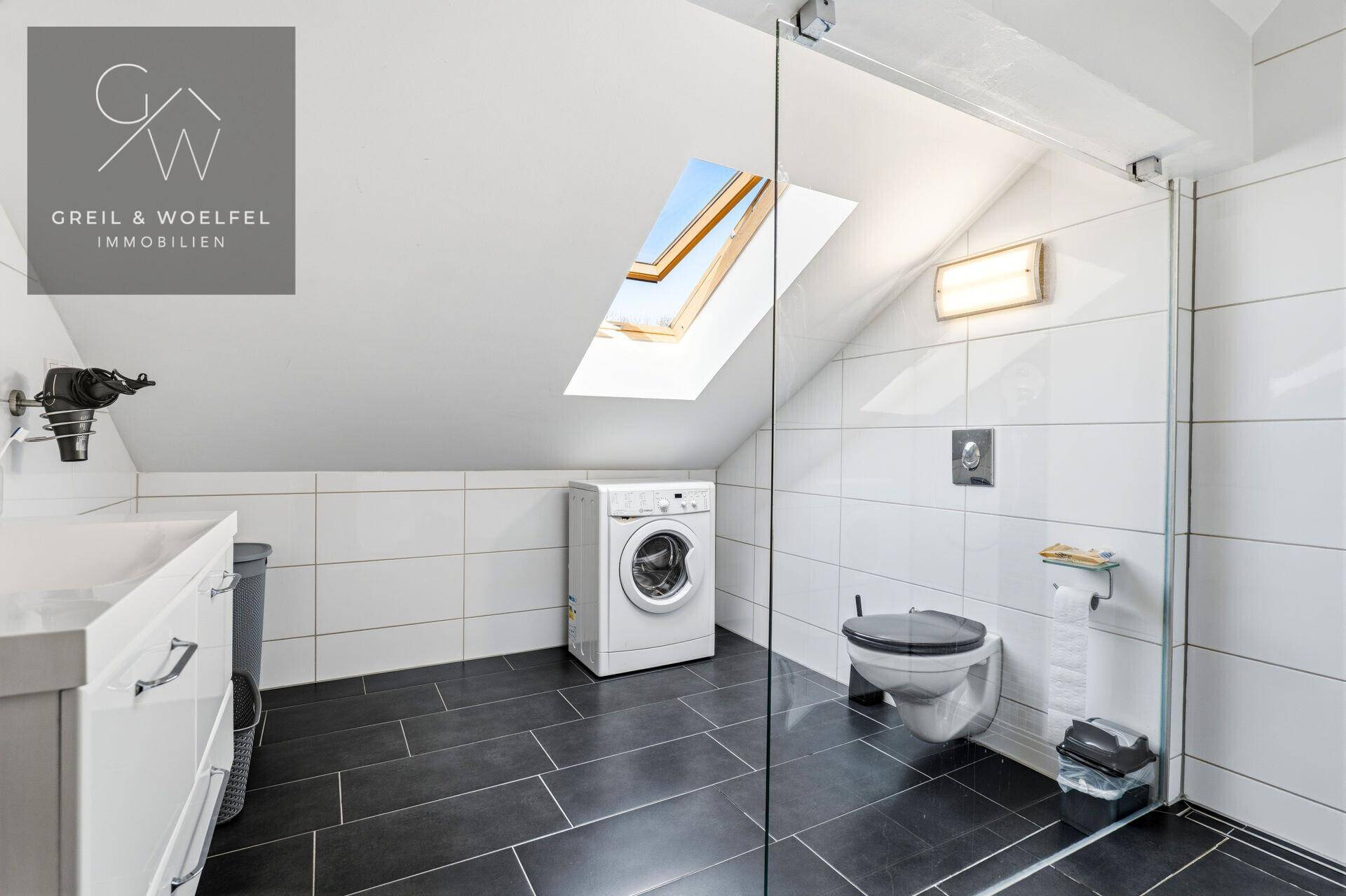 Modernes Badezimmer mit dunklen Bodenfliesen, Glasdusche, Toilette und Dachfenster für viel Licht.