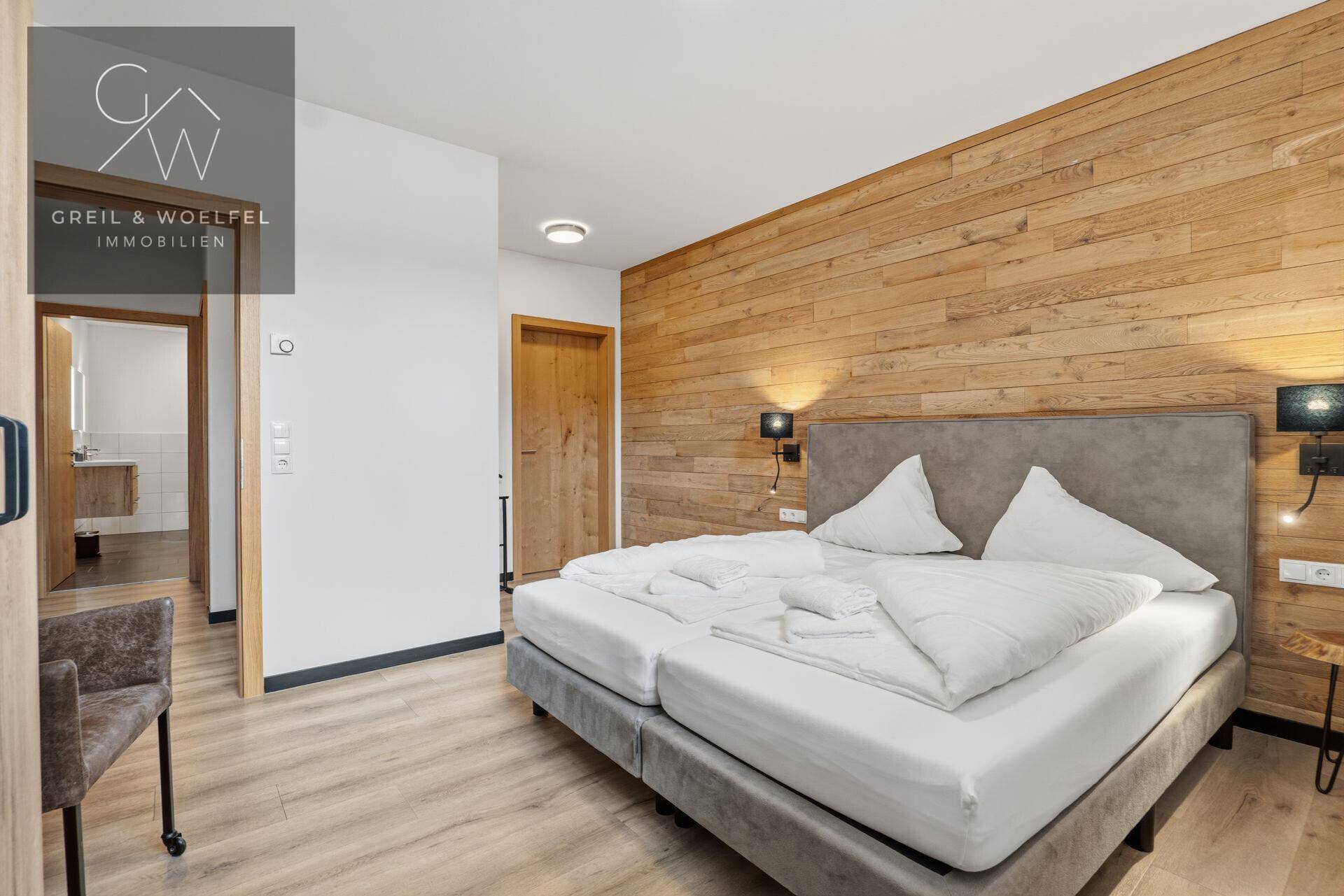Gemütliches Schlafzimmer mit Holzwand, Doppelbett und Blick in ein modernes Badezimmer.