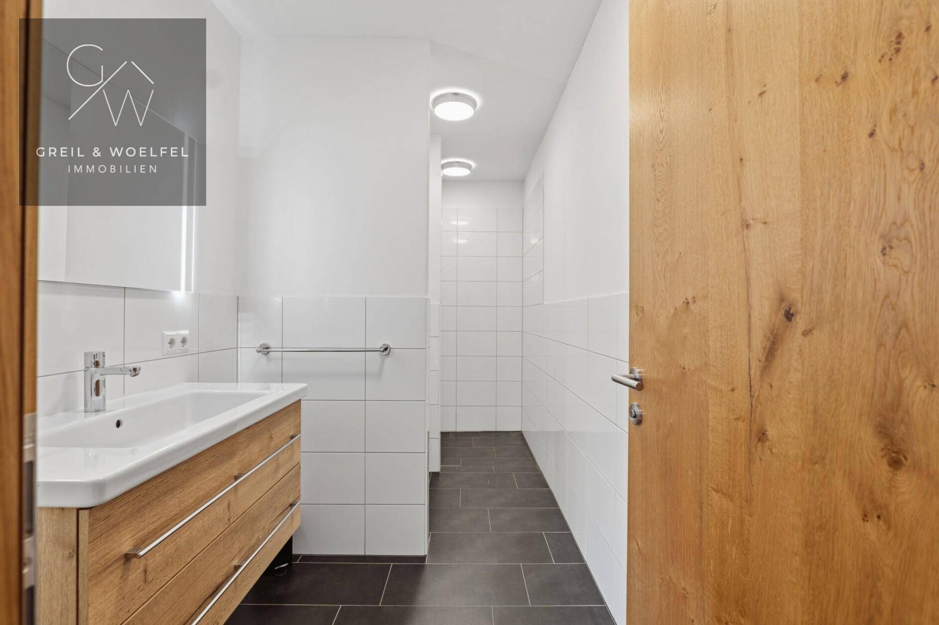 Modernes Badezimmer mit weiß gefliesten Wänden, dunklem Boden und Holzwaschtisch.