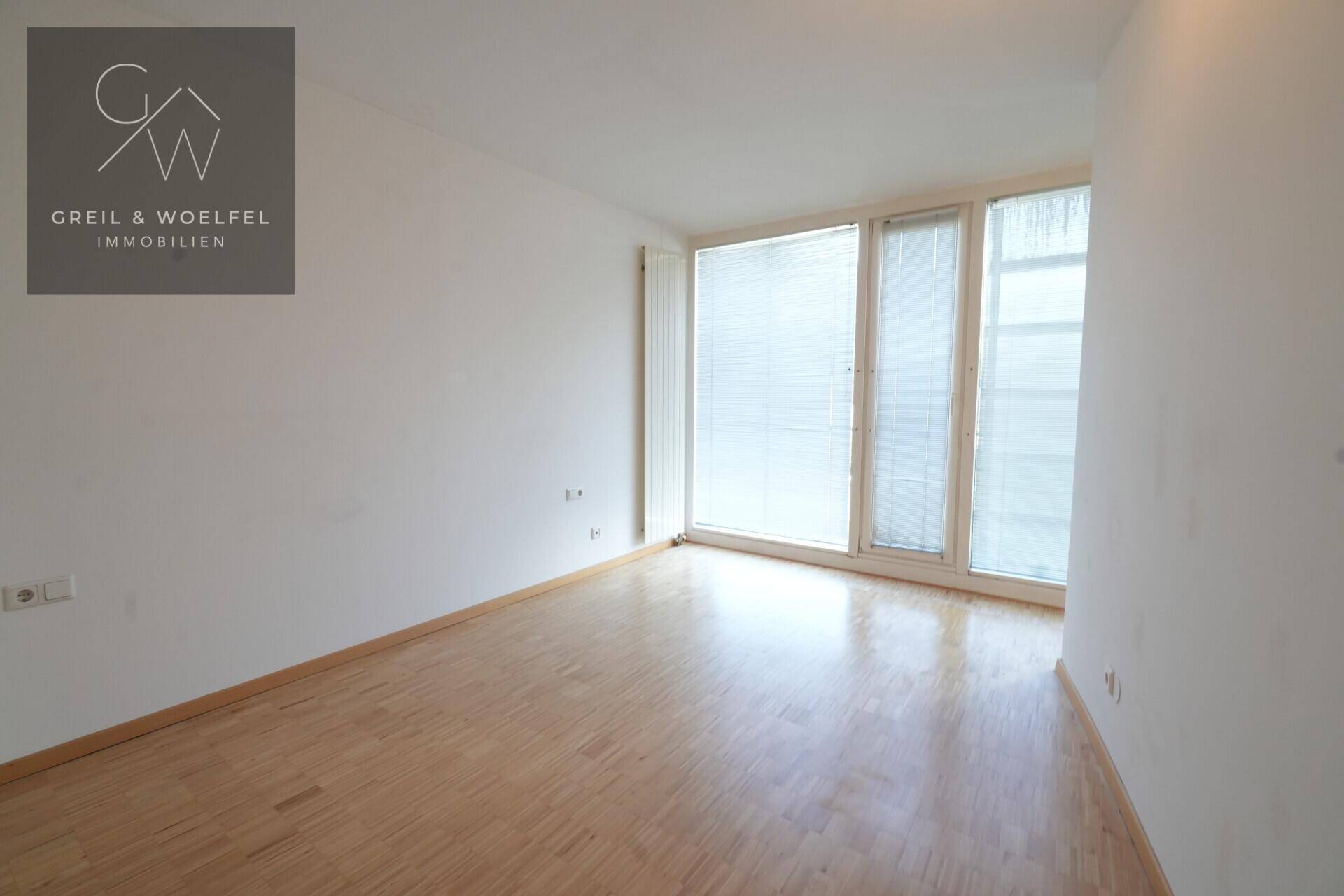 Leeres Schlafzimmer mit Parkettboden, großen Fenstern und einem Heizkörper.
