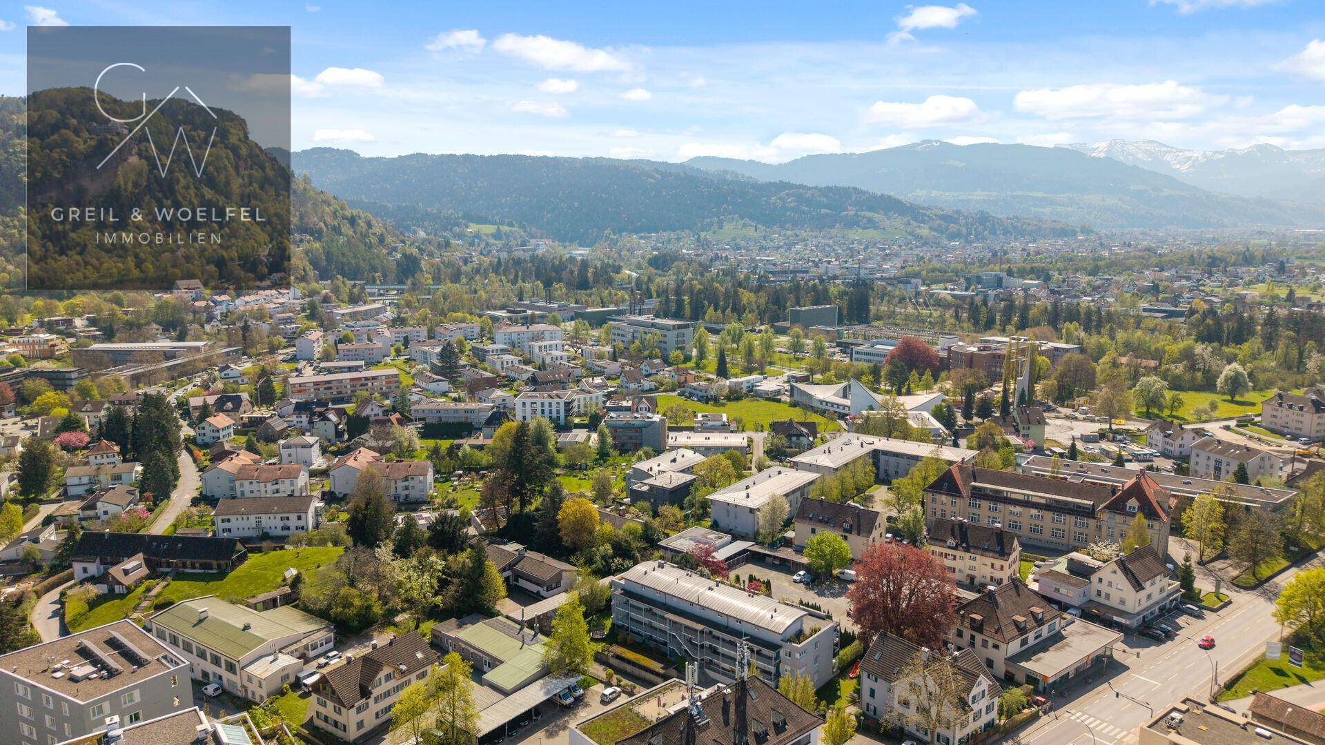 Weitwinkelaufnahme einer Stadtlandschaft mit grünen Hügeln und Bergen am Horizont.