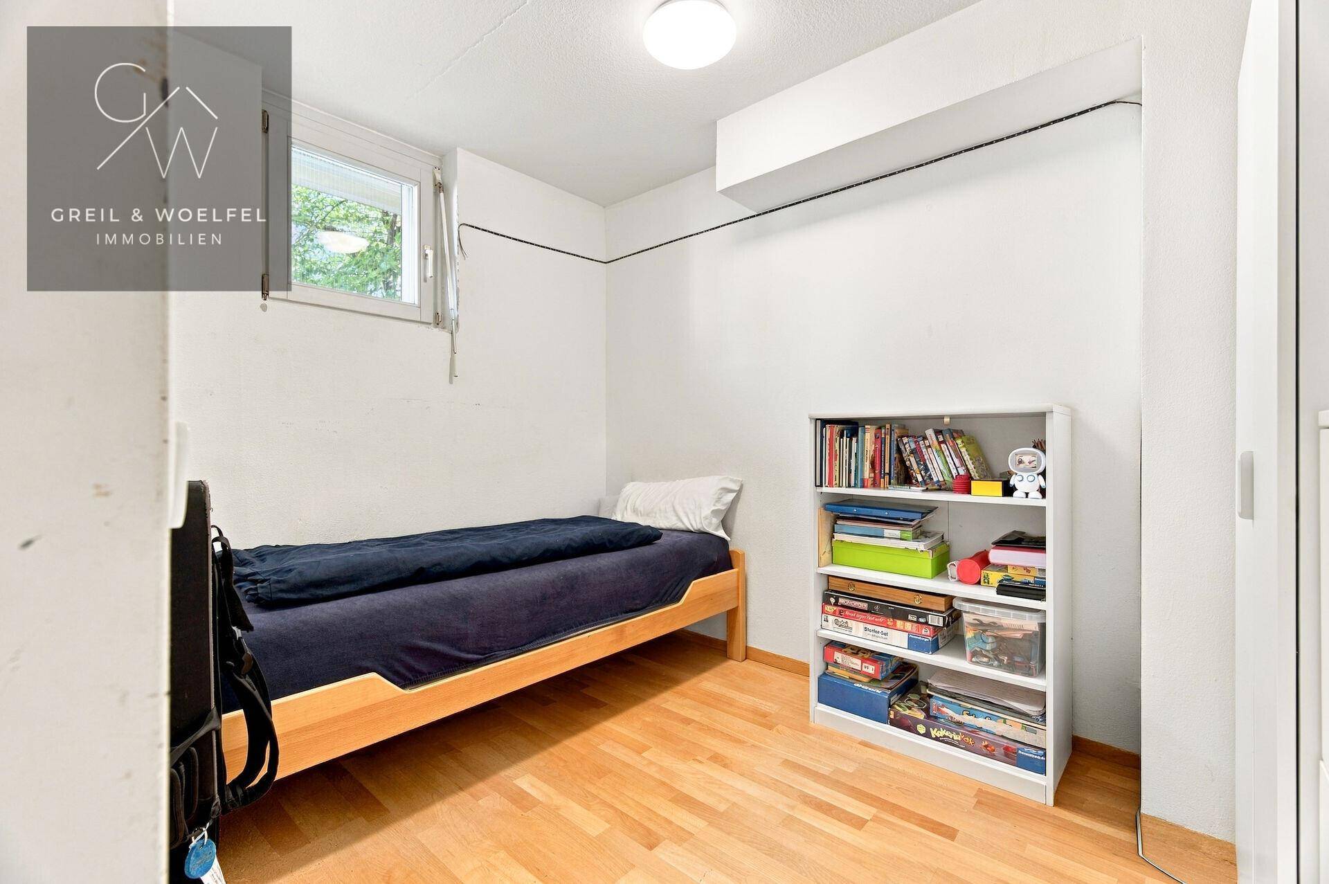Helles Zimmer mit Einzelbett, Bücherregal und Fenster für ausreichend Tageslicht.