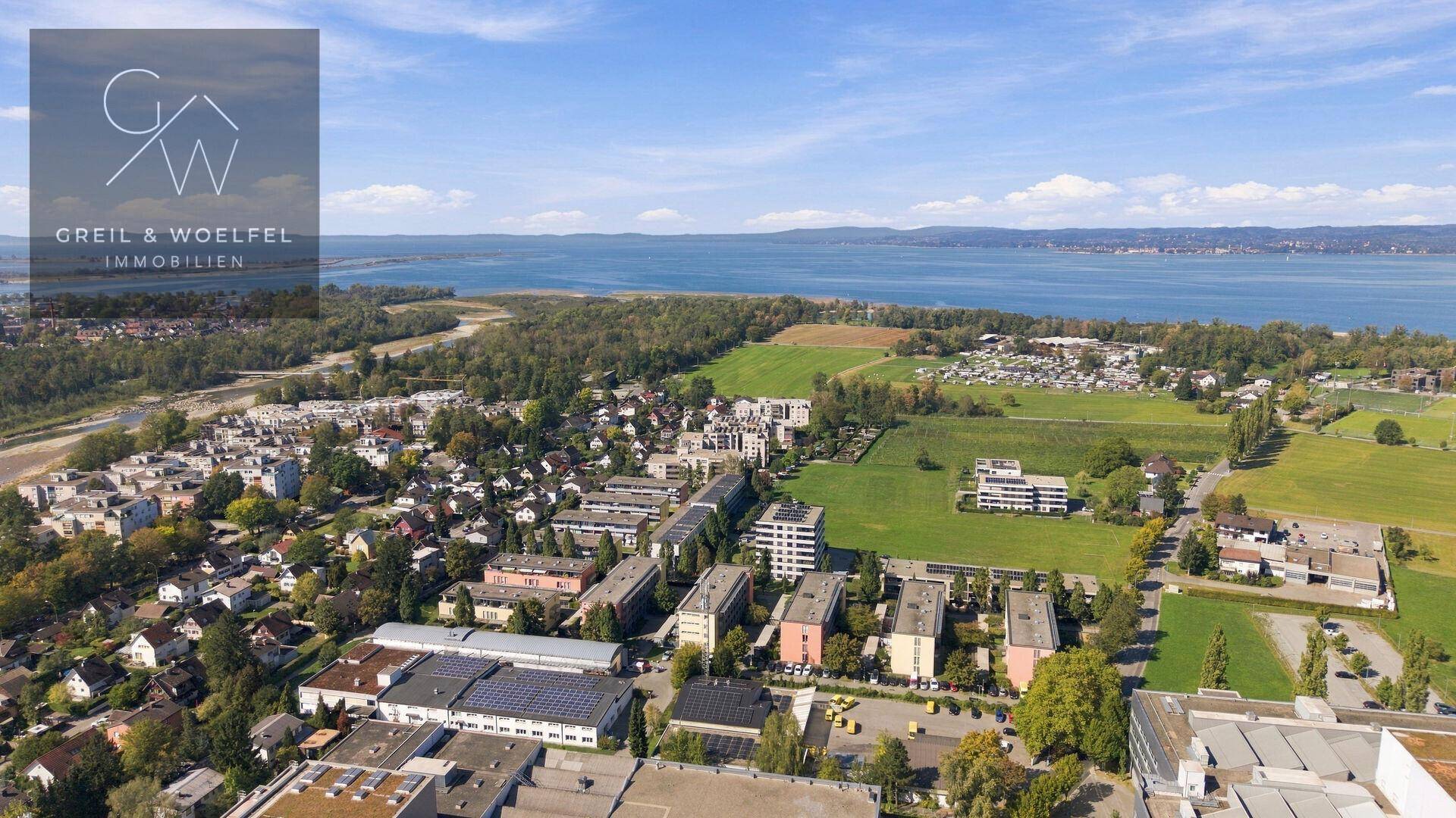 Weitläufige Luftaufnahme einer Siedlung am Seeufer mit grünen Feldern und Bergen im Hintergrund.