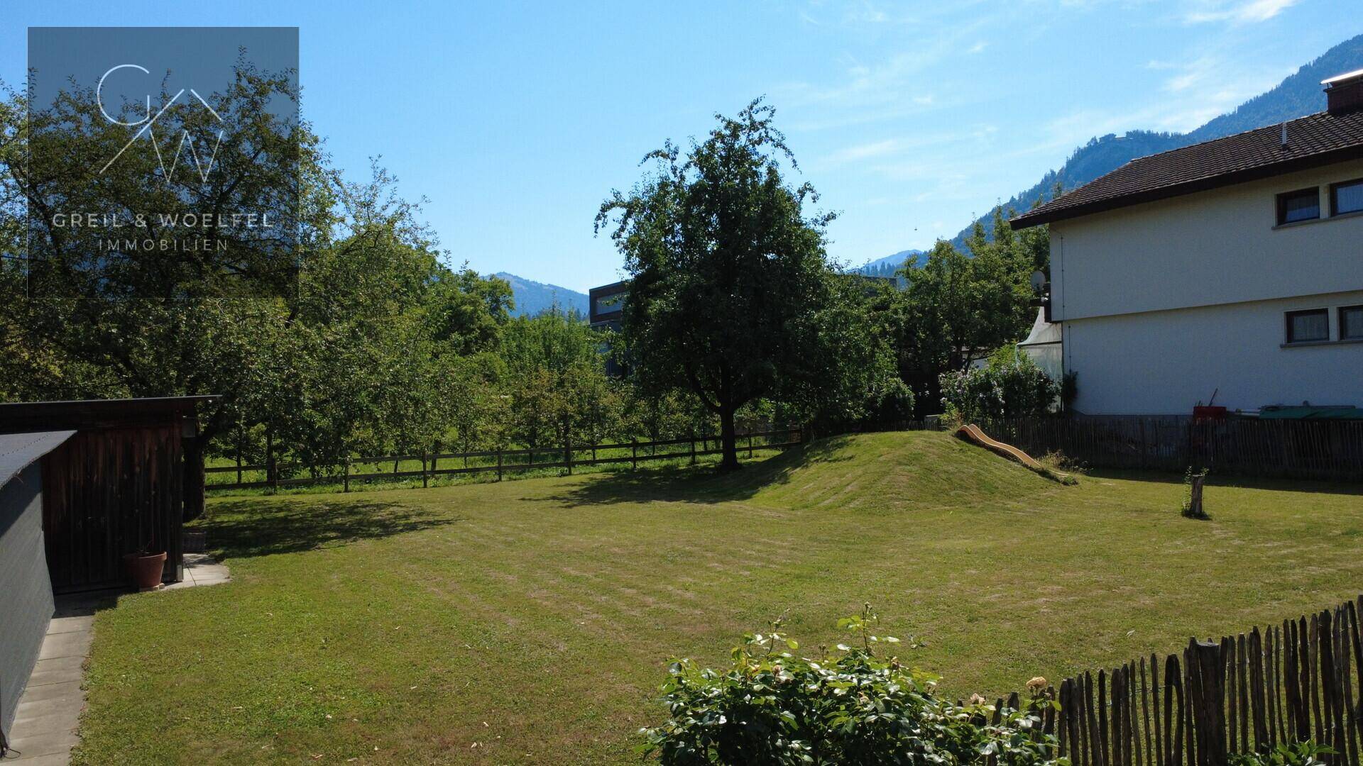 Großer grüner Garten mit Rasenfläche, Bäumen, einem kleinen Hügel und Bergblick im Hintergrund.