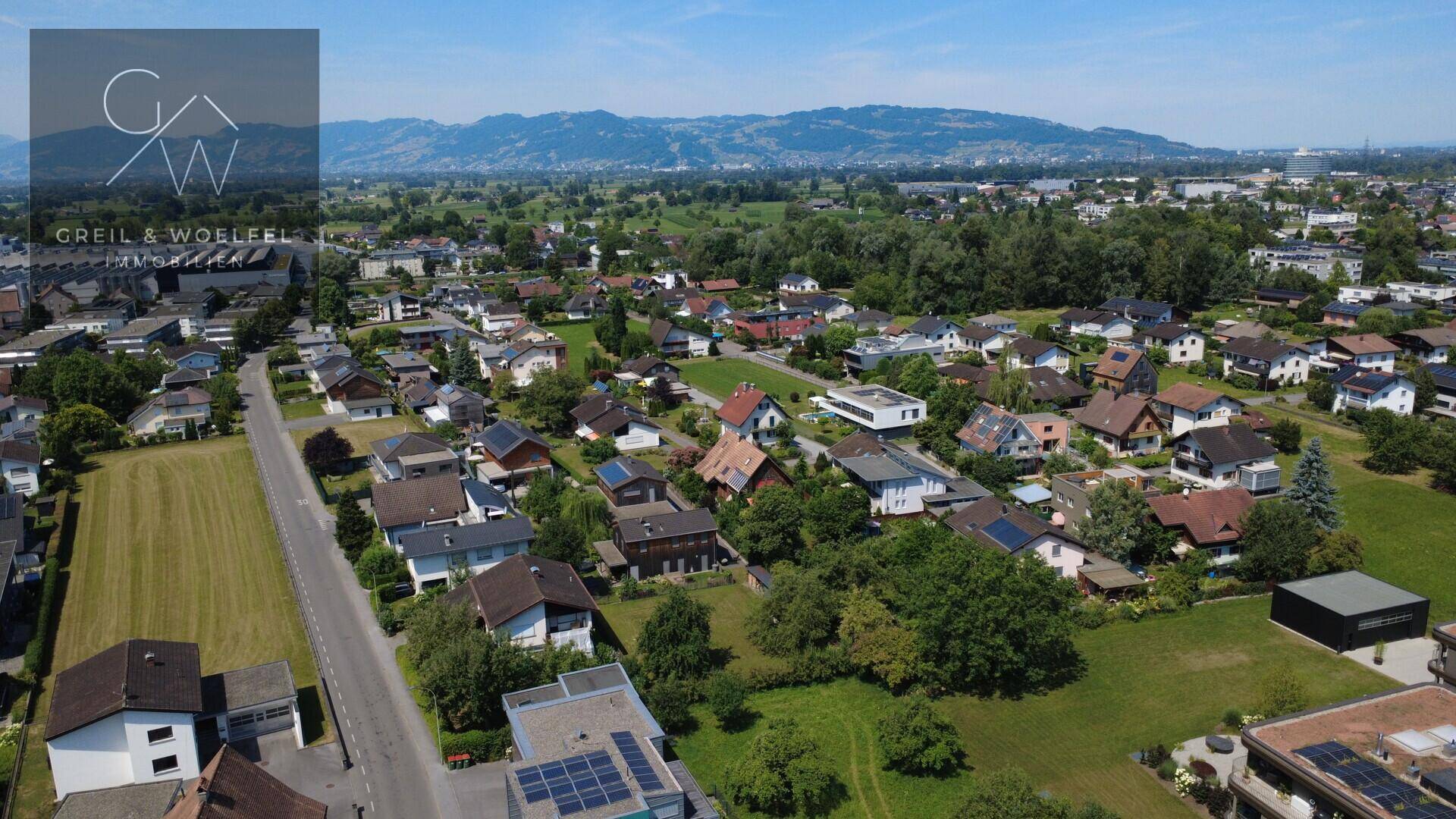 Luftaufnahme eines Wohngebiets mit Häusern, grünen Feldern und Bergen im Hintergrund, wobei ein Grundstück hervorgehoben ist.