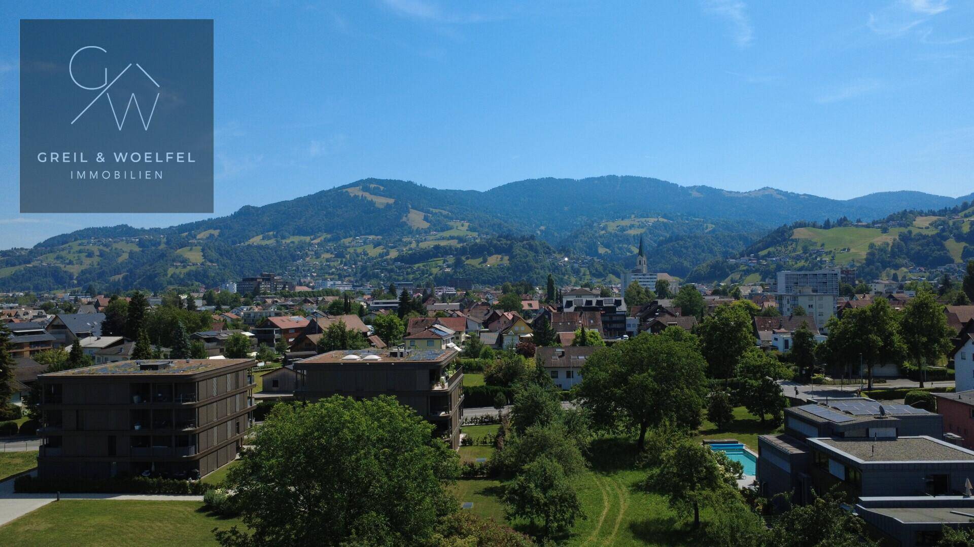 Luftaufnahme einer Stadt oder eines Dorfes mit vielen Häusern, Grünflächen und Bergen im Hintergrund.