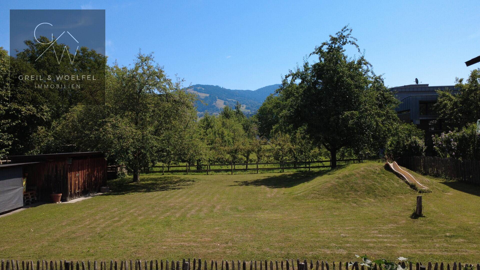 Weitläufiger grüner Garten mit Rasen, Bäumen, einer Rutsche auf einem Hügel und Bergblick im Hintergrund.