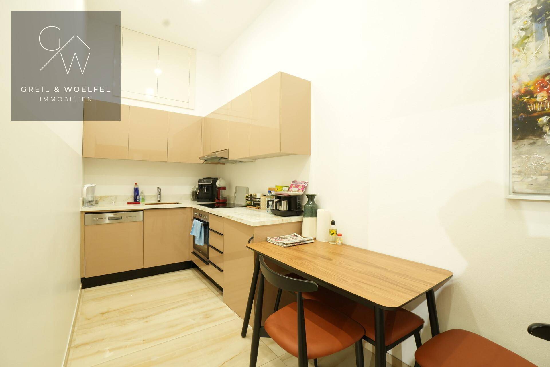 Kompakte Teeküche mit beigen Schränken, Spülmaschine und kleinem Esstisch.