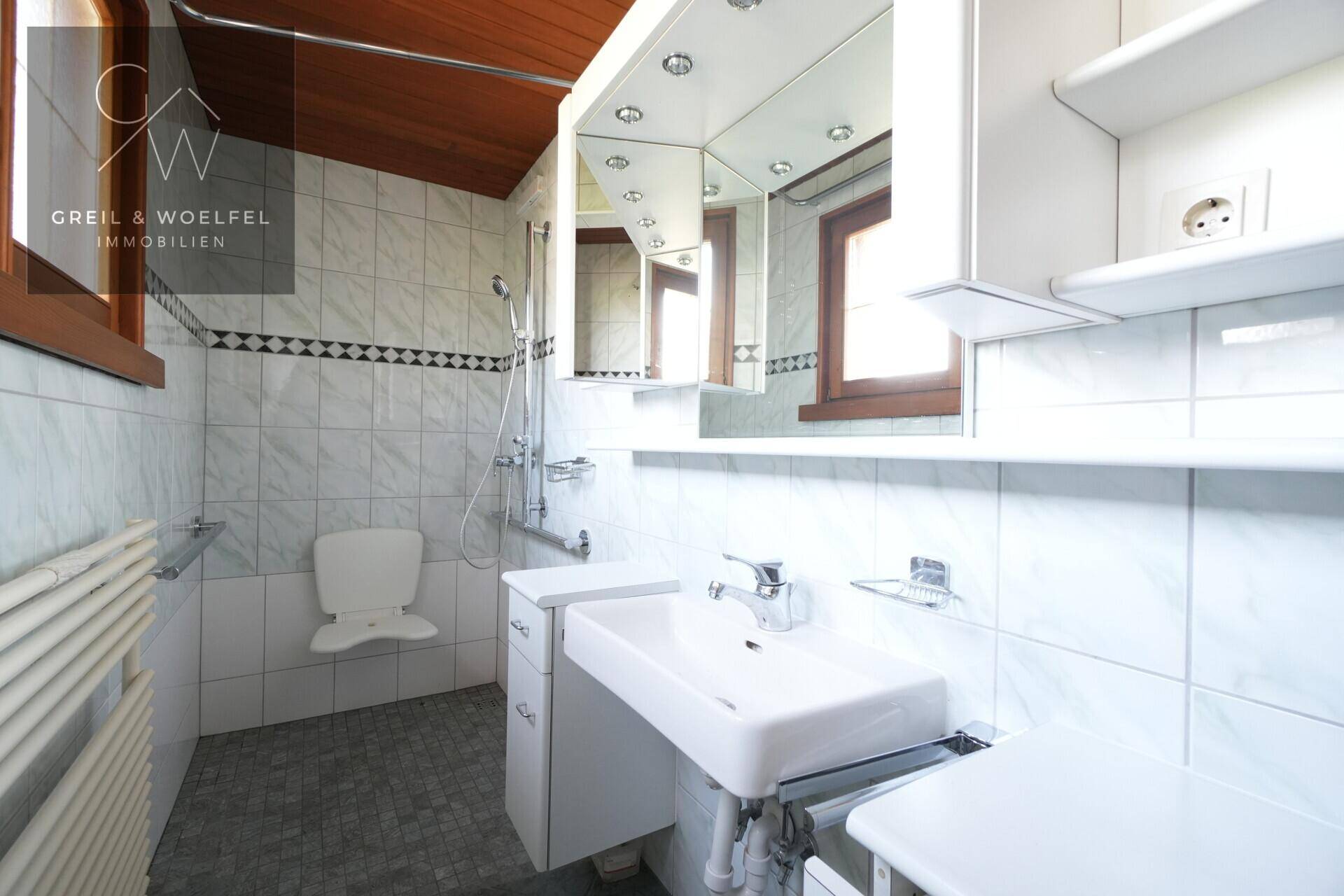 Modernisiertes Badezimmer mit ebenerdiger Dusche, Haltegriffen und Fenster, barrierefrei gestaltet.