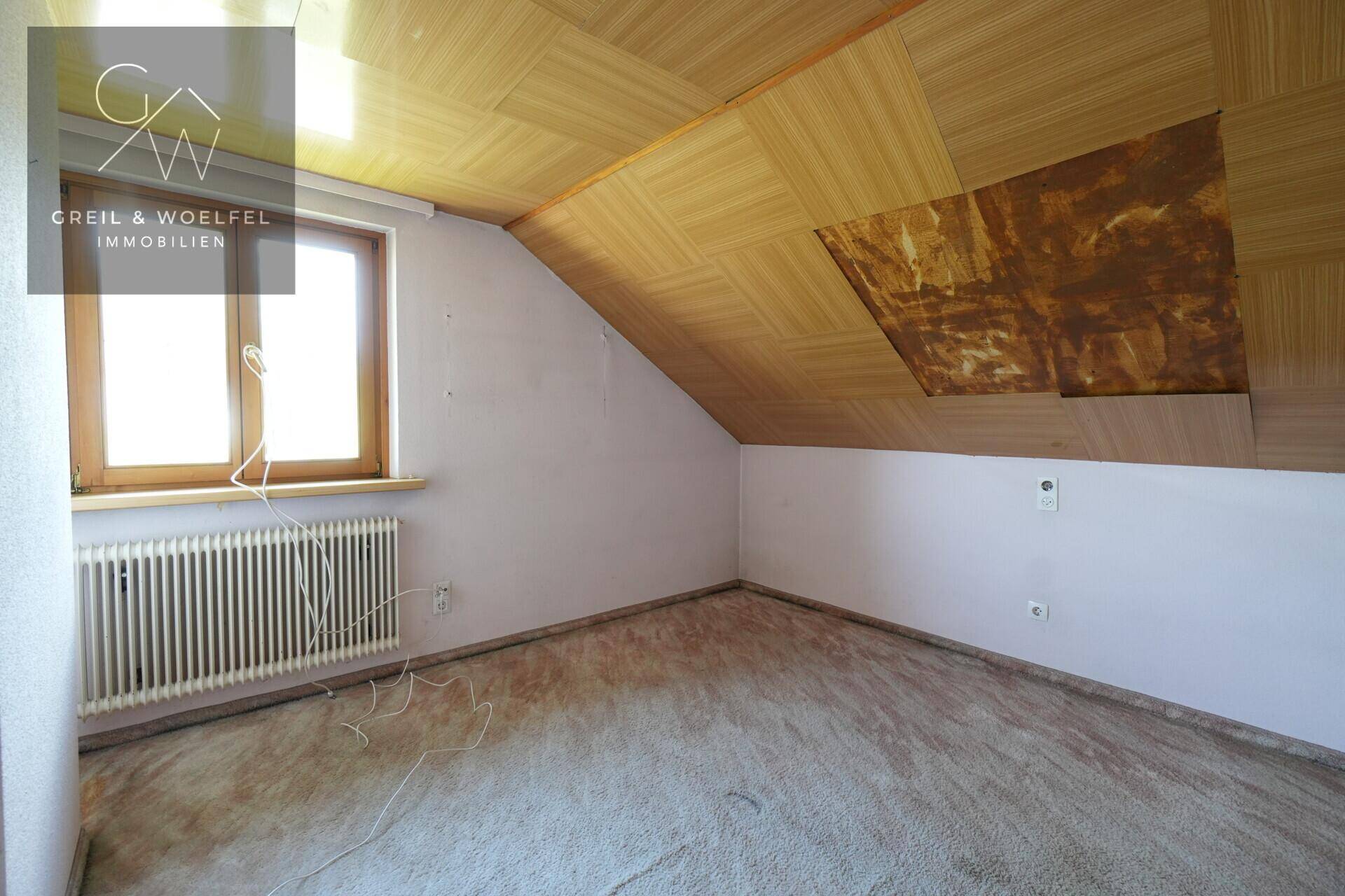 Dachgeschosszimmer mit schrägen Wänden, Teppichboden und Fenster, bereit zur Renovierung.