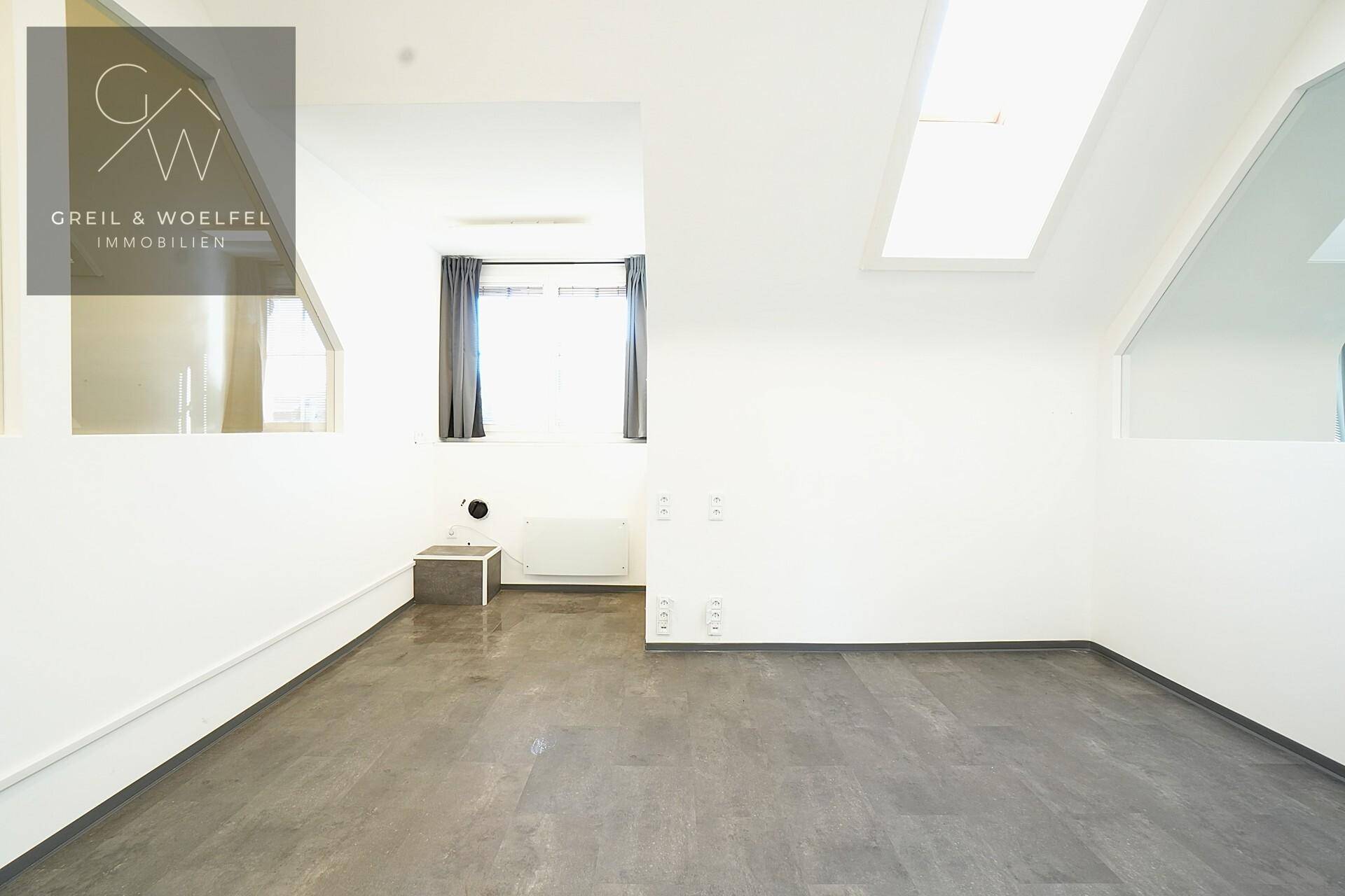 Leerer Büroraum mit Dachschräge, Fenster und dunklem Vinylboden.