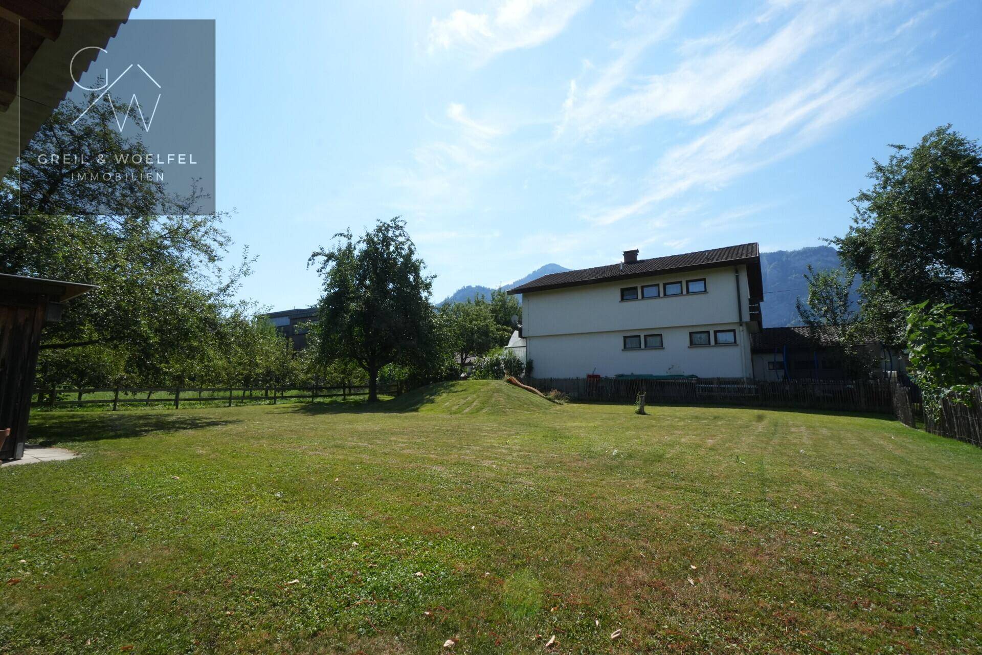 Großer grüner Garten mit Rasenfläche, Bäumen, einem kleinen Hügel und einem Haus im Hintergrund mit Bergblick.