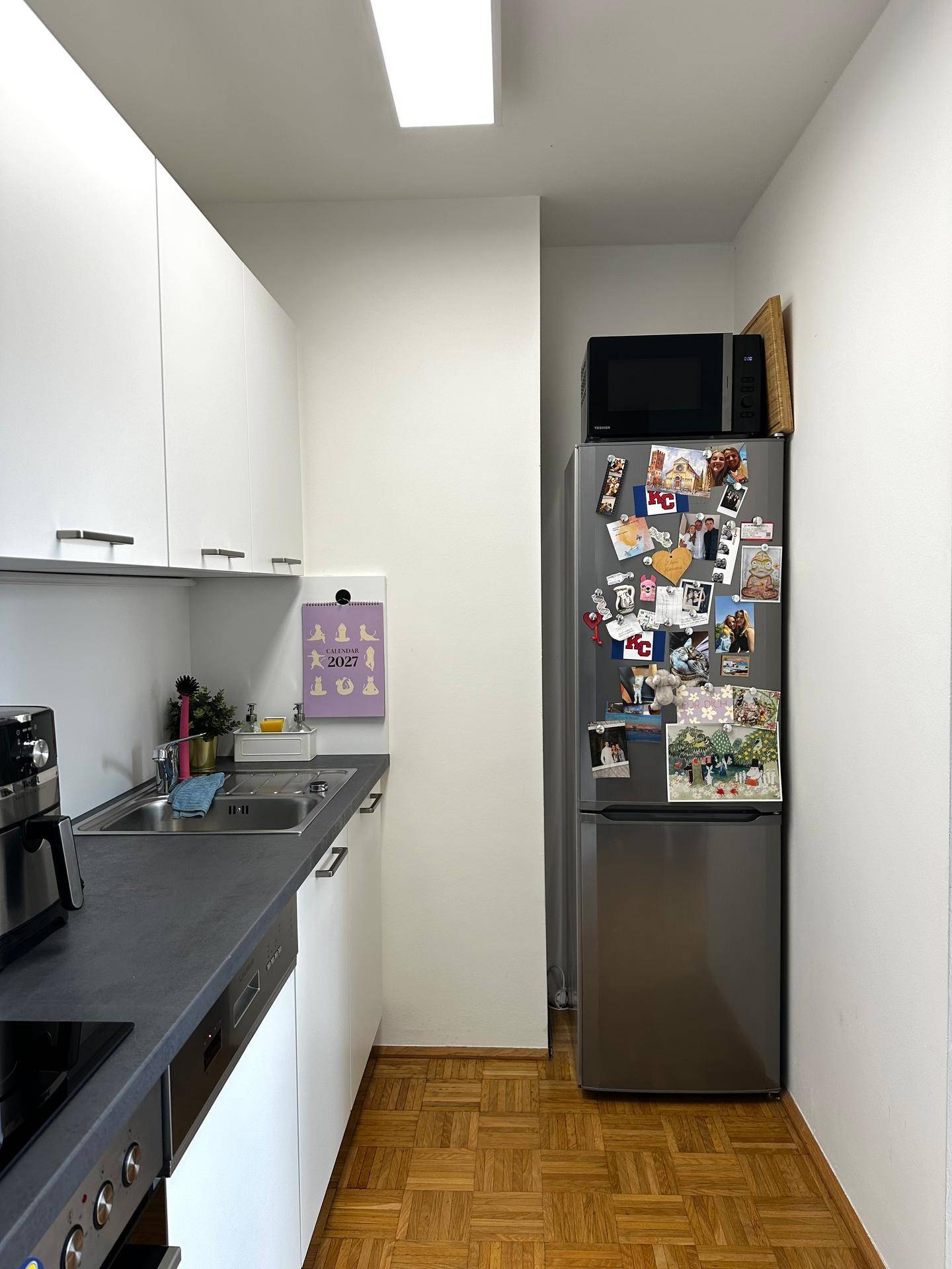 Schmale Küche mit weißen Oberschränken, Spüle und einem Kühlschrank mit Mikrowelle.