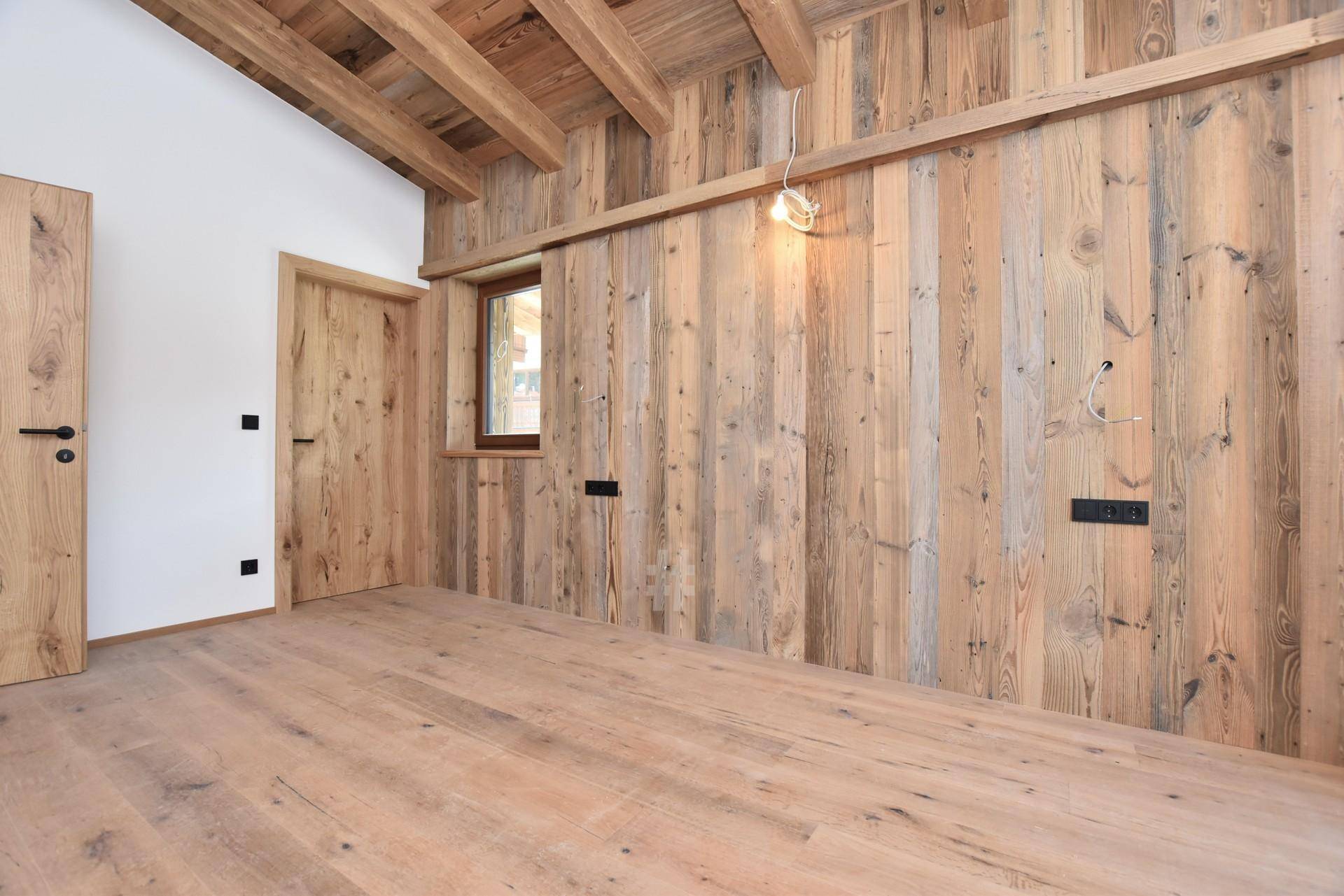 Raum mit Holzboden, Holzwandverkleidung und sichtbaren Holzbalken, mit einer Holztür und kleinem Fenster.