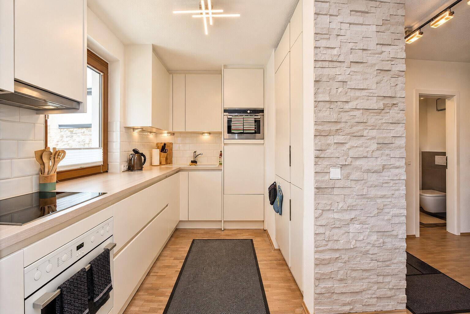 Moderne weiße Küche mit Einbaugeräten, Fenster und dekorativer Steinwandverkleidung.