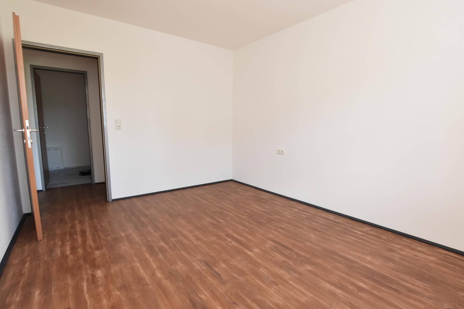 Leerer Raum mit Laminatboden und einer offenen Tür, die zu einem weiteren Zimmer führt.