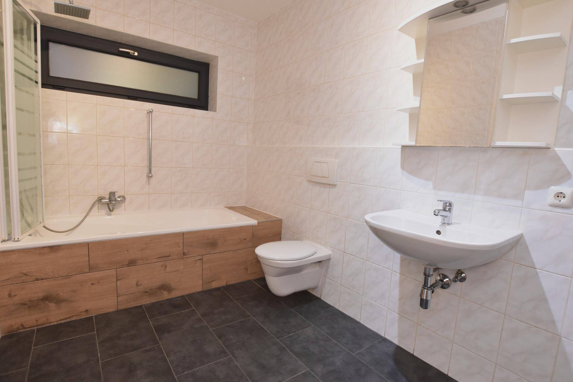 Helles Badezimmer mit Badewanne, WC, Waschbecken und einem schmalen Fenster.