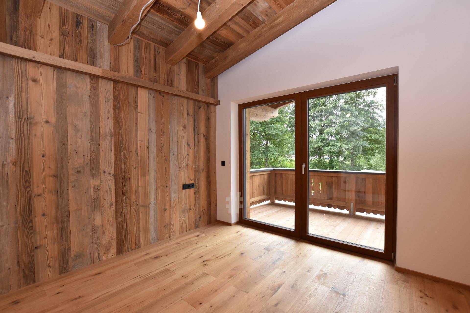 Raum mit Holzboden, Holzwandverkleidung und Zugang zu einem Balkon mit traditionellem Holzgeländer.