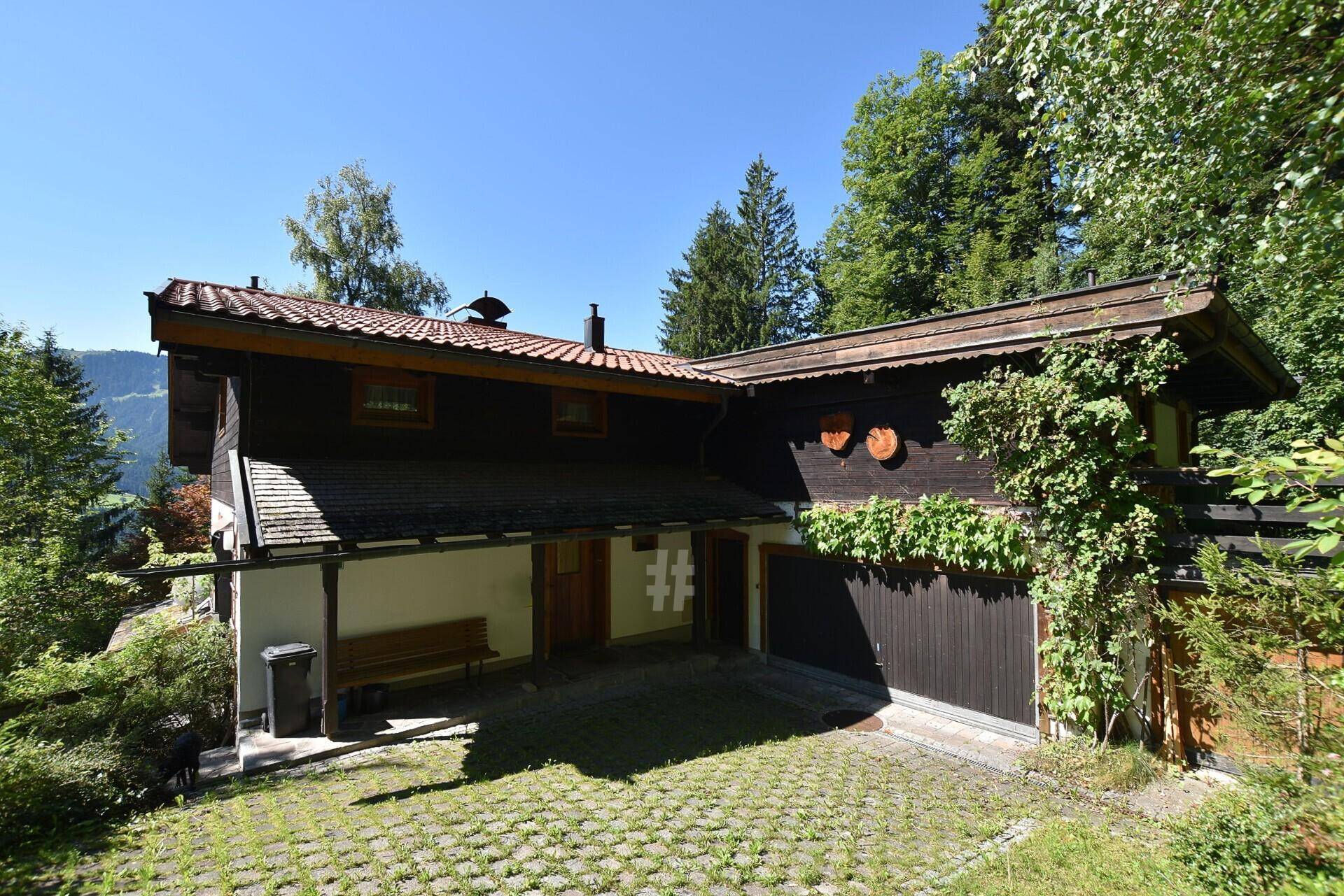 Seitenansicht des Hauses mit Holzfassade, Garage und gepflastertem Vorplatz umgeben von Bäumen.