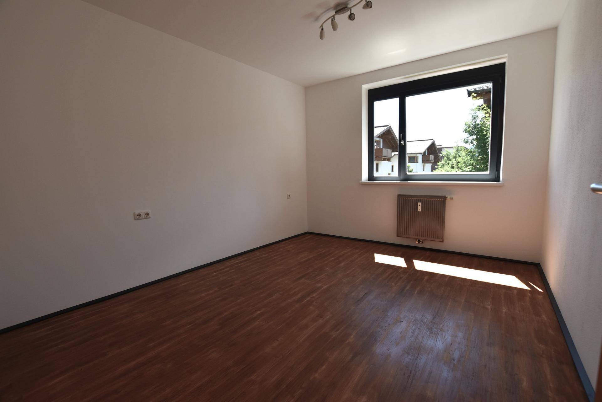 Heller, leerer Raum mit großem Fenster und warmem Laminatboden, der viel Tageslicht hereinlässt.