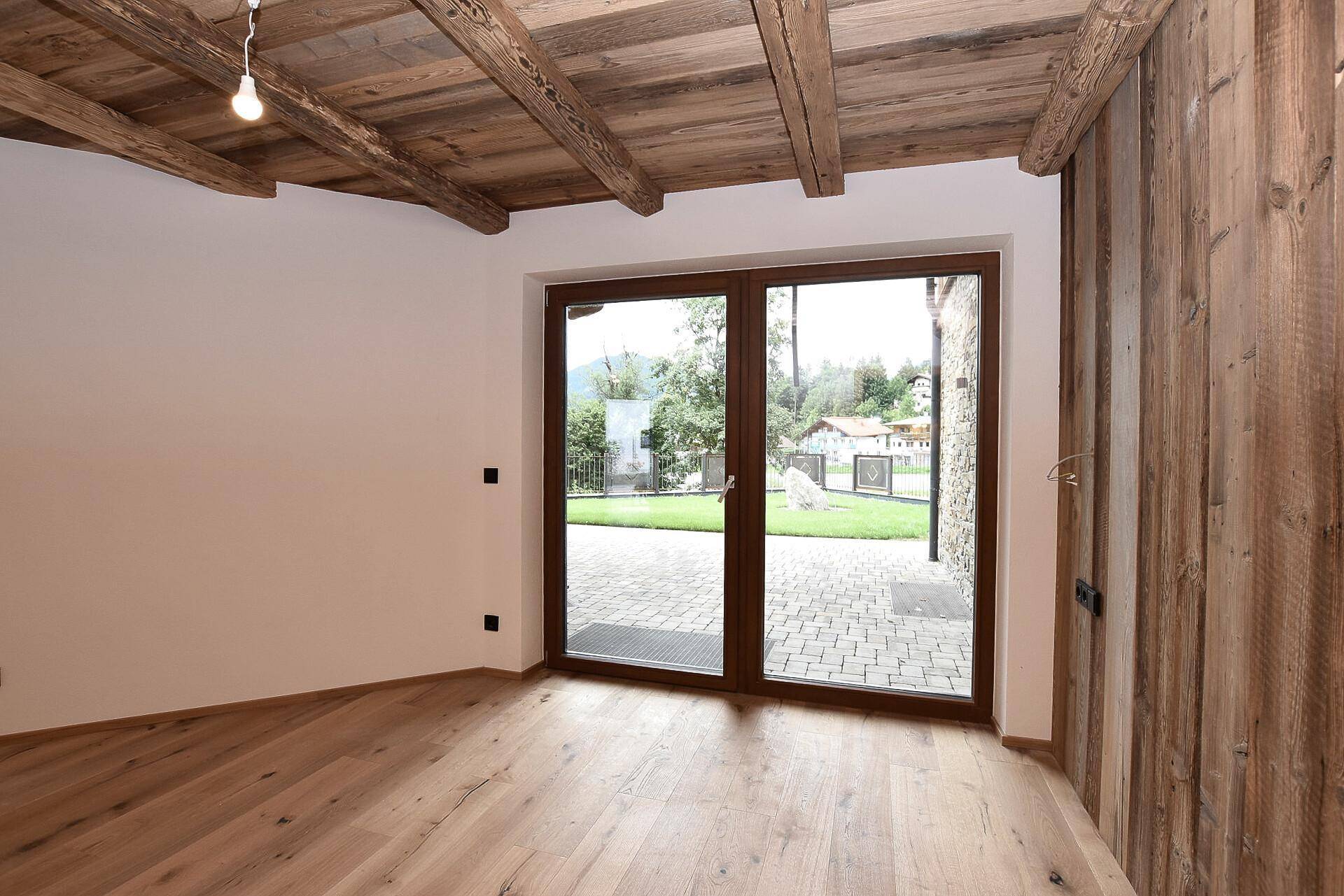 Gemütlicher Raum mit Holzboden, sichtbaren Holzbalken und Zugang zu einer gepflasterten Terrasse.