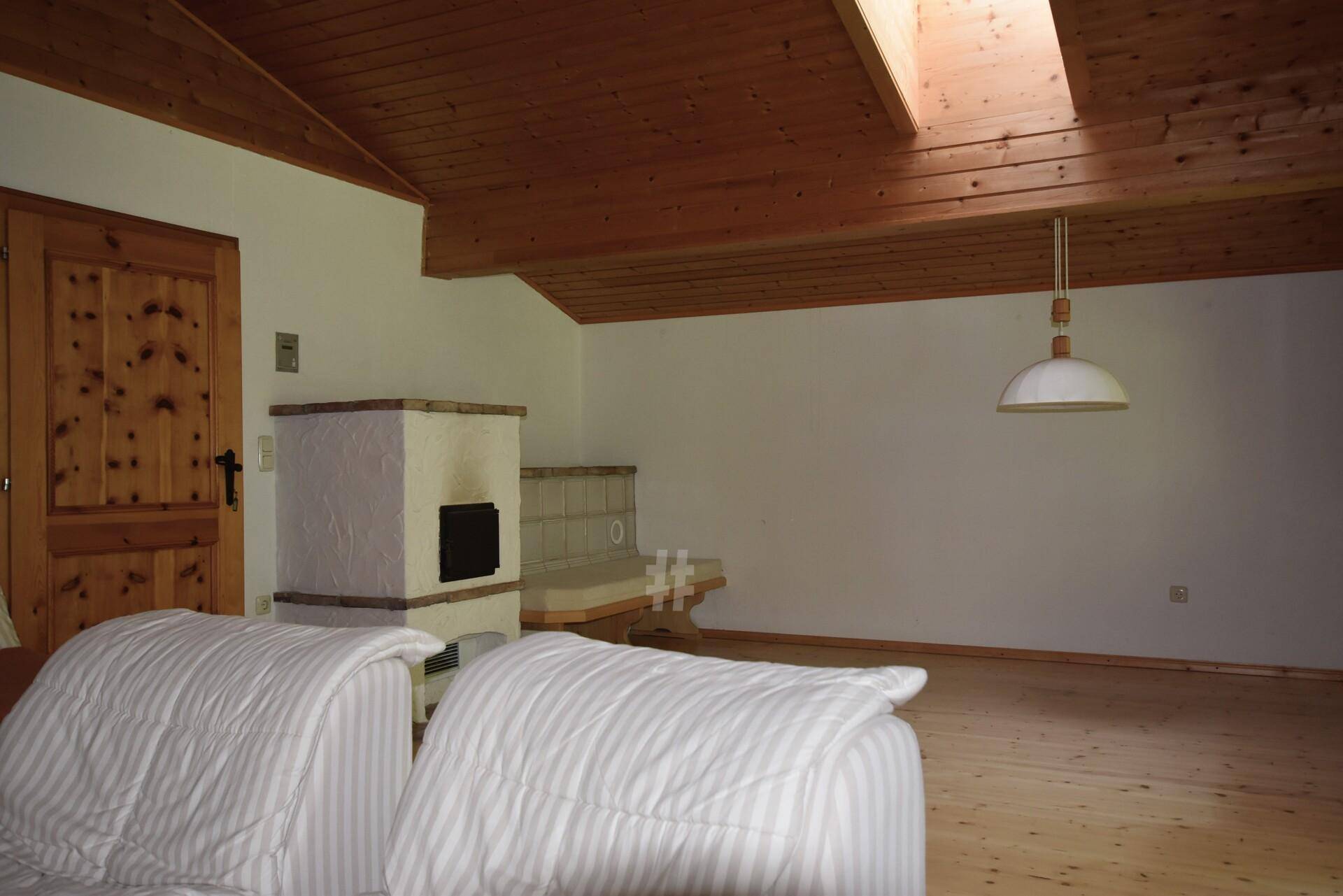 Helles Zimmer im Dachgeschoss mit Holzbalkendecke, Kachelofen und einem Dachfenster für natürliches Licht.