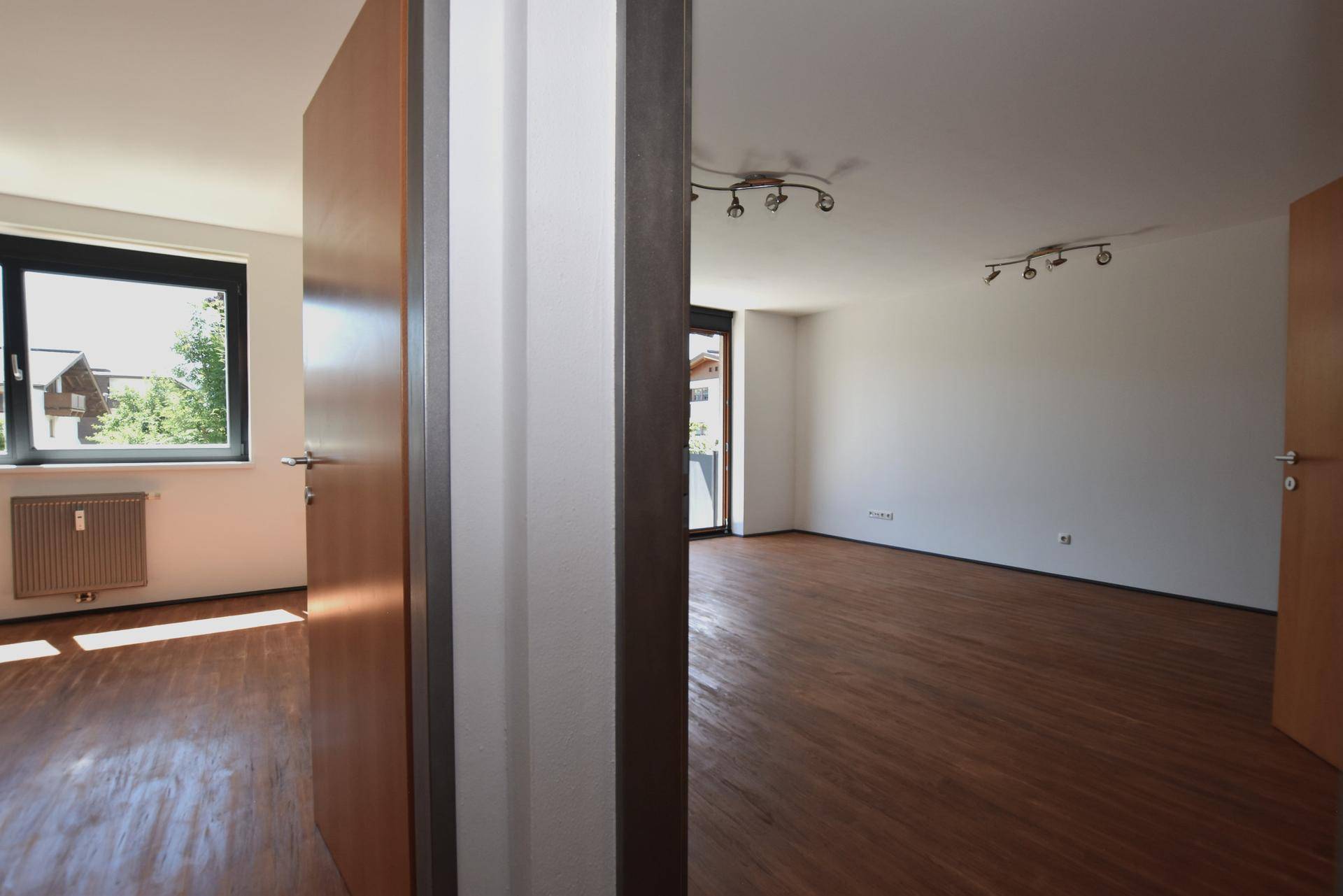 Blick durch eine Tür in zwei helle, leere Räume mit Laminatboden und Fenstern.