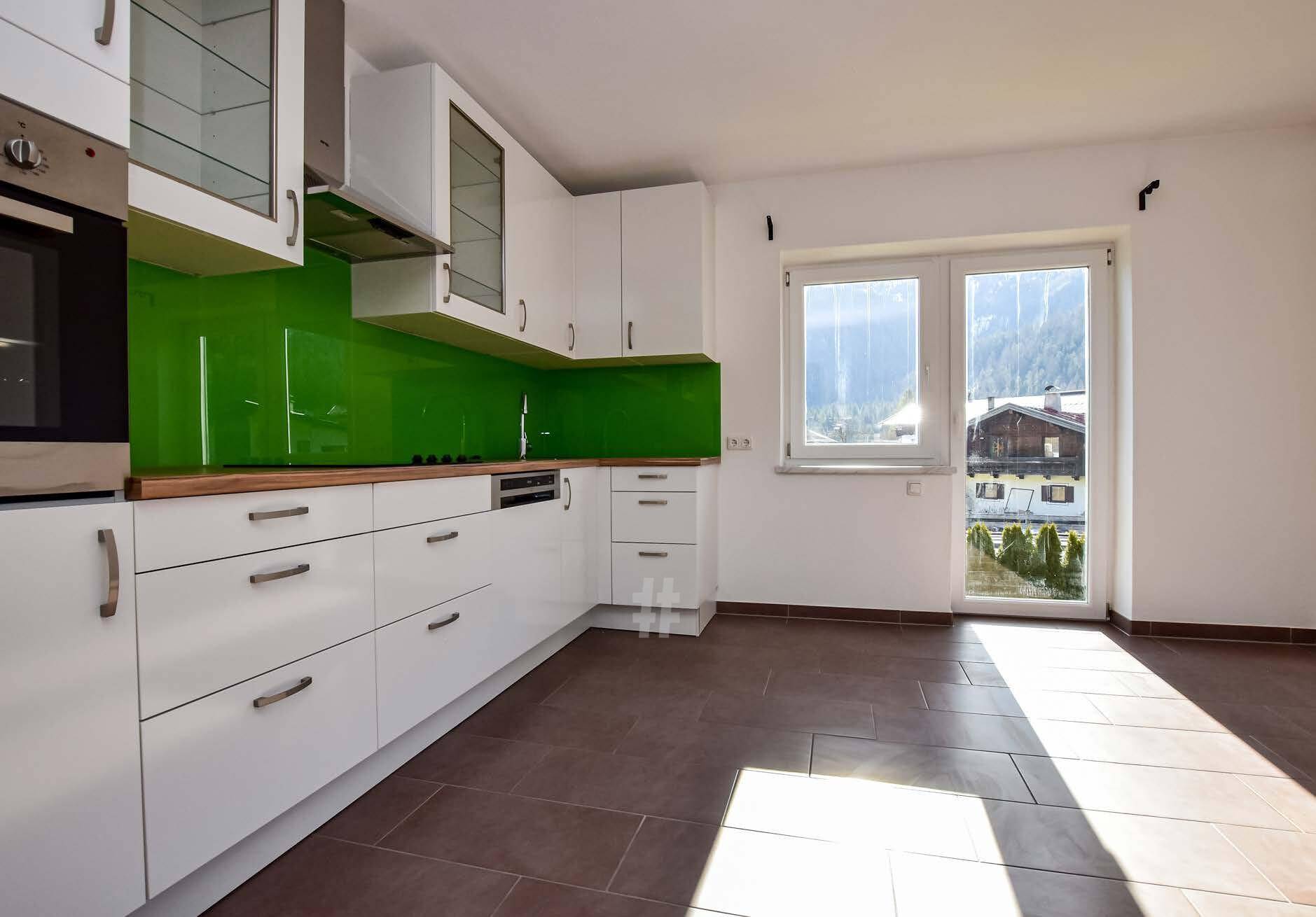 Helle Küche mit weißen Schränken, grüner Rückwand und Zugang nach draußen mit Bergblick.