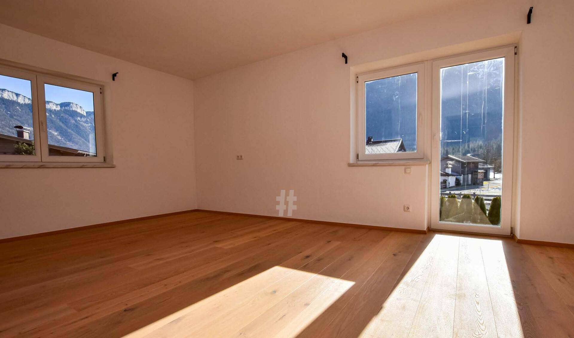 Lichtdurchfluteter Raum mit Holzboden, Fenstern und Zugang nach draußen, mit Bergblick.