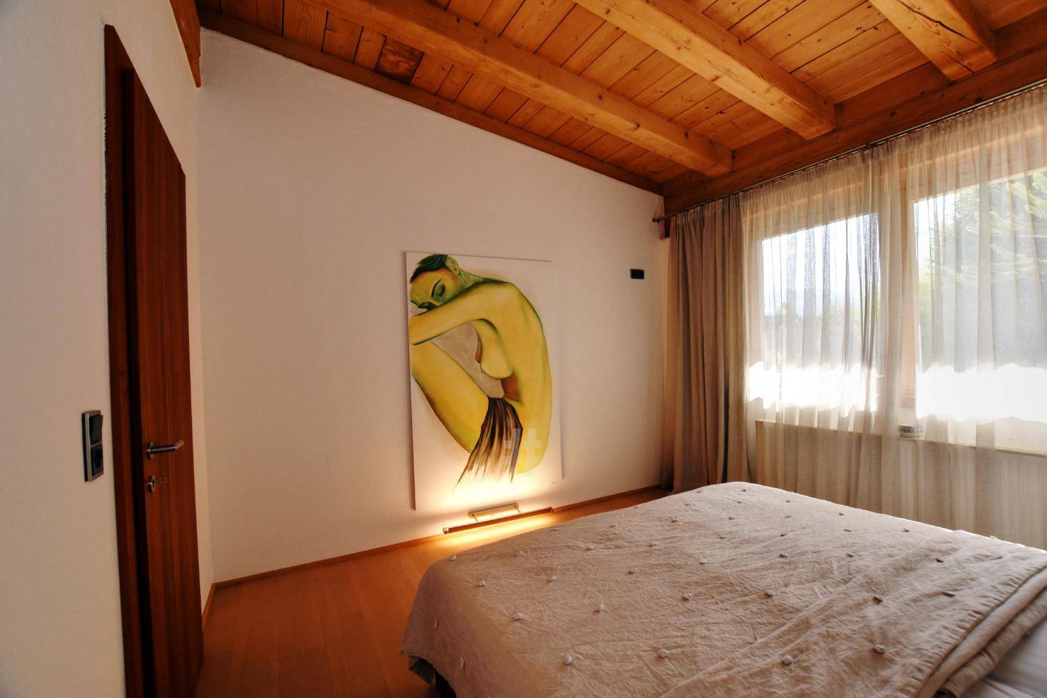 Gemütliches Schlafzimmer mit Holzbalkendecke, Parkettboden und großem Fenster.