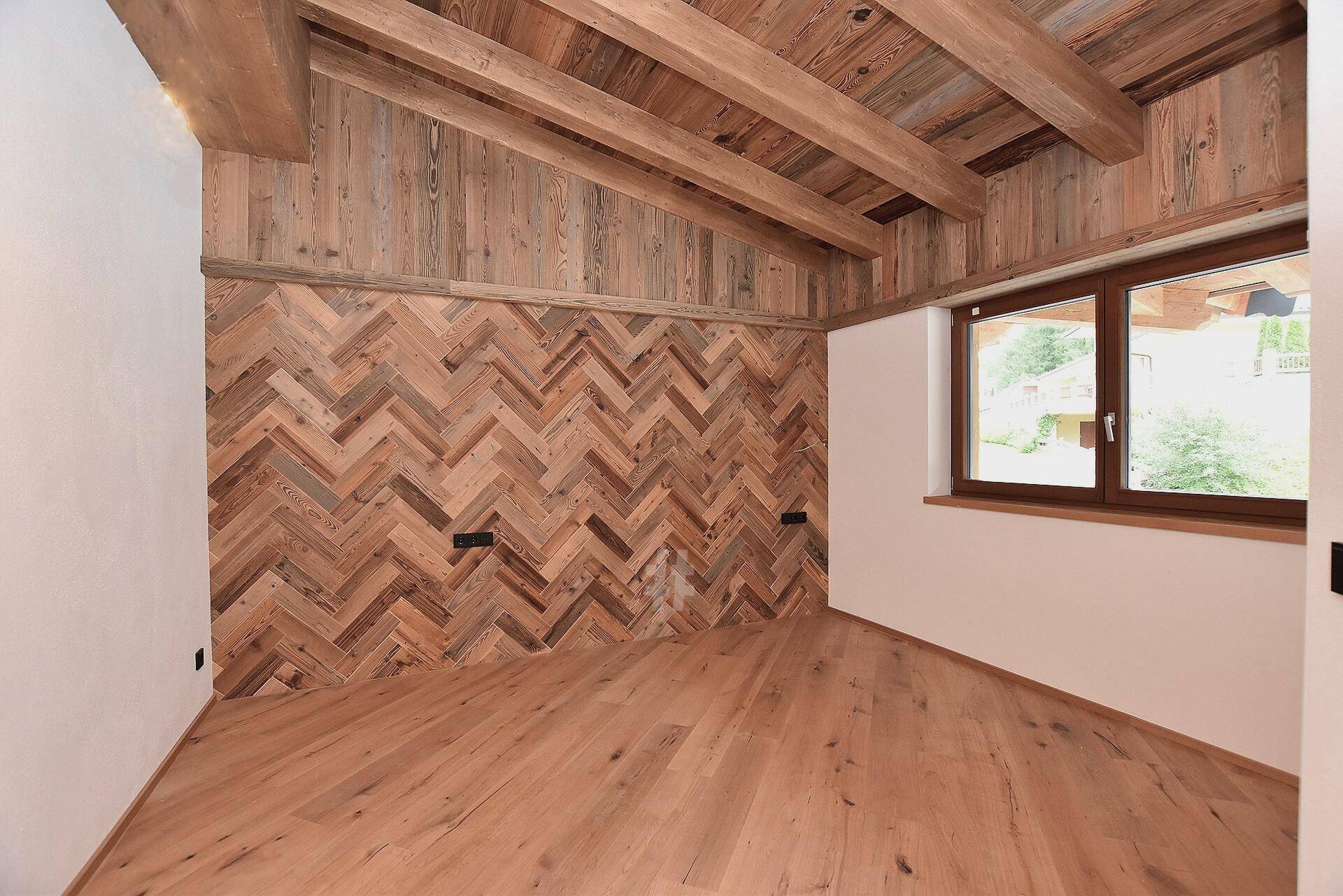 Raum mit Holzboden, Holzbalkendecke und einer dekorativen Holzwand im Fischgrätmuster.