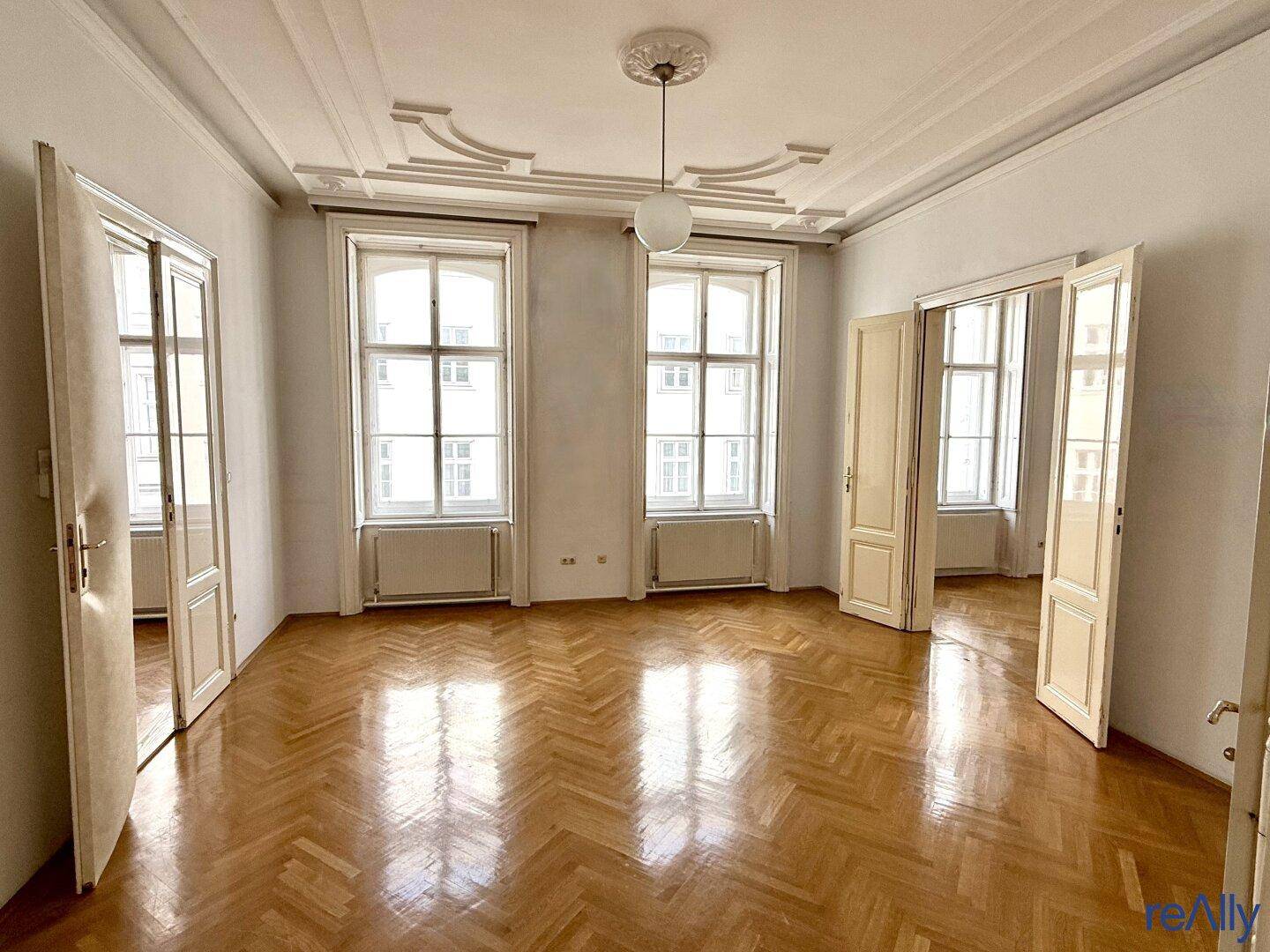 Geräumiges Wohnzimmer mit Fischgrätparkett, hohen Decken und Stuckverzierungen, hell durch große Fenster.