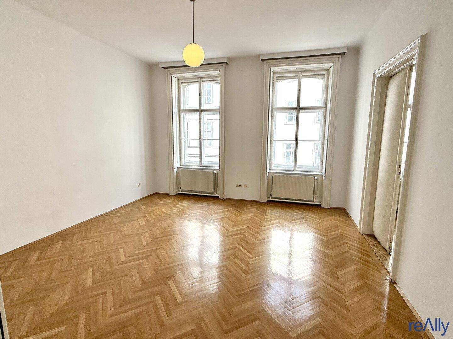 Heller Raum mit Fischgrätparkett und zwei hohen Fenstern, ideal für verschiedene Nutzungen.