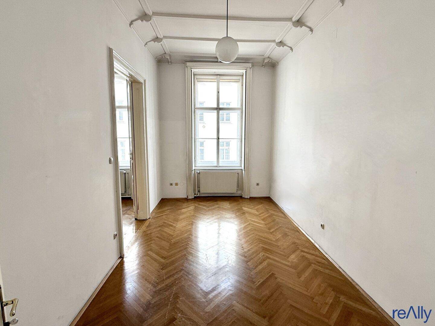 Heller Raum mit Fischgrätparkett und einem hohen Fenster, ideal für verschiedene Nutzungen.