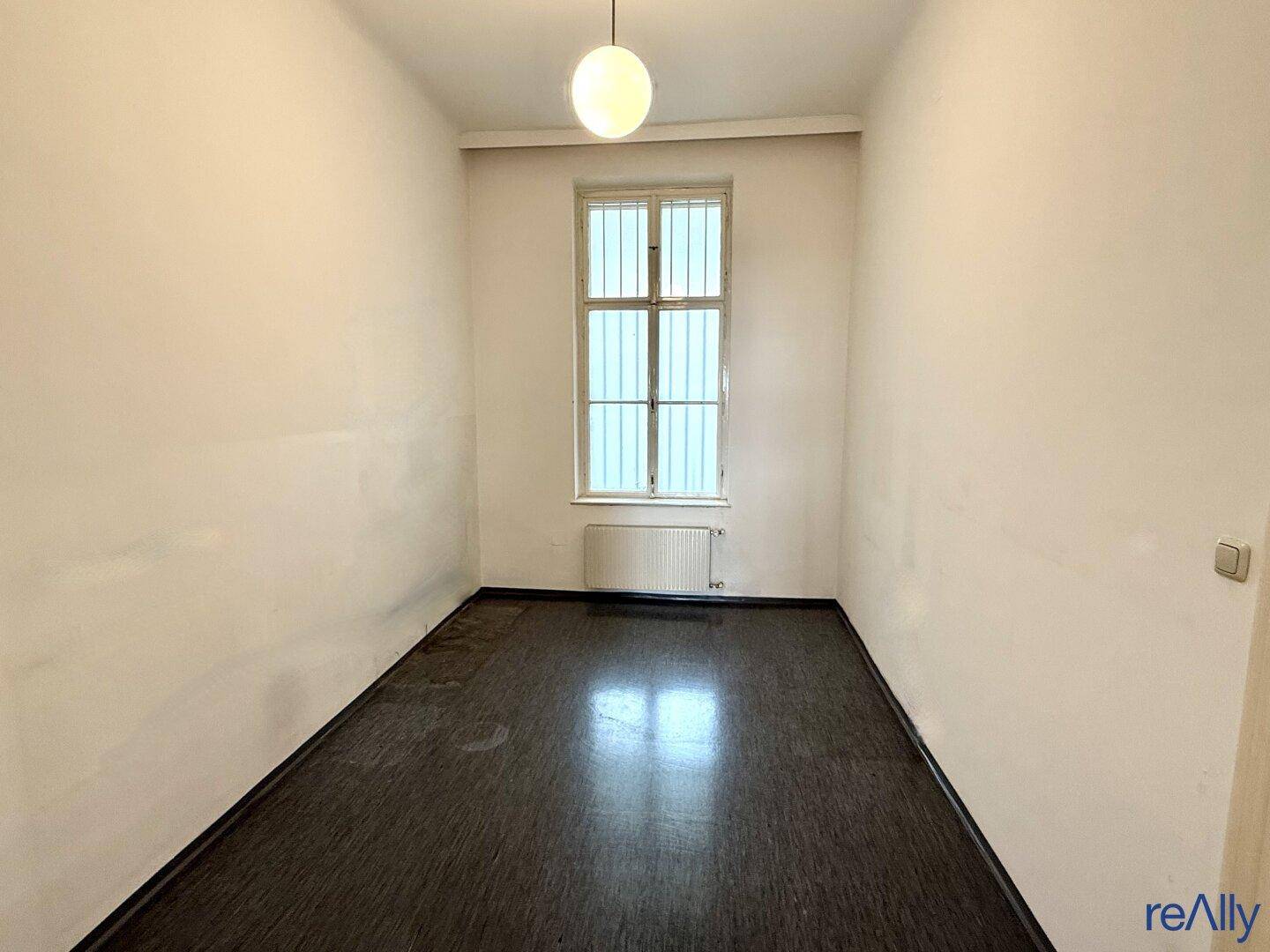 Kleiner, schmaler Raum mit dunklem Boden und einem vergitterten Fenster.