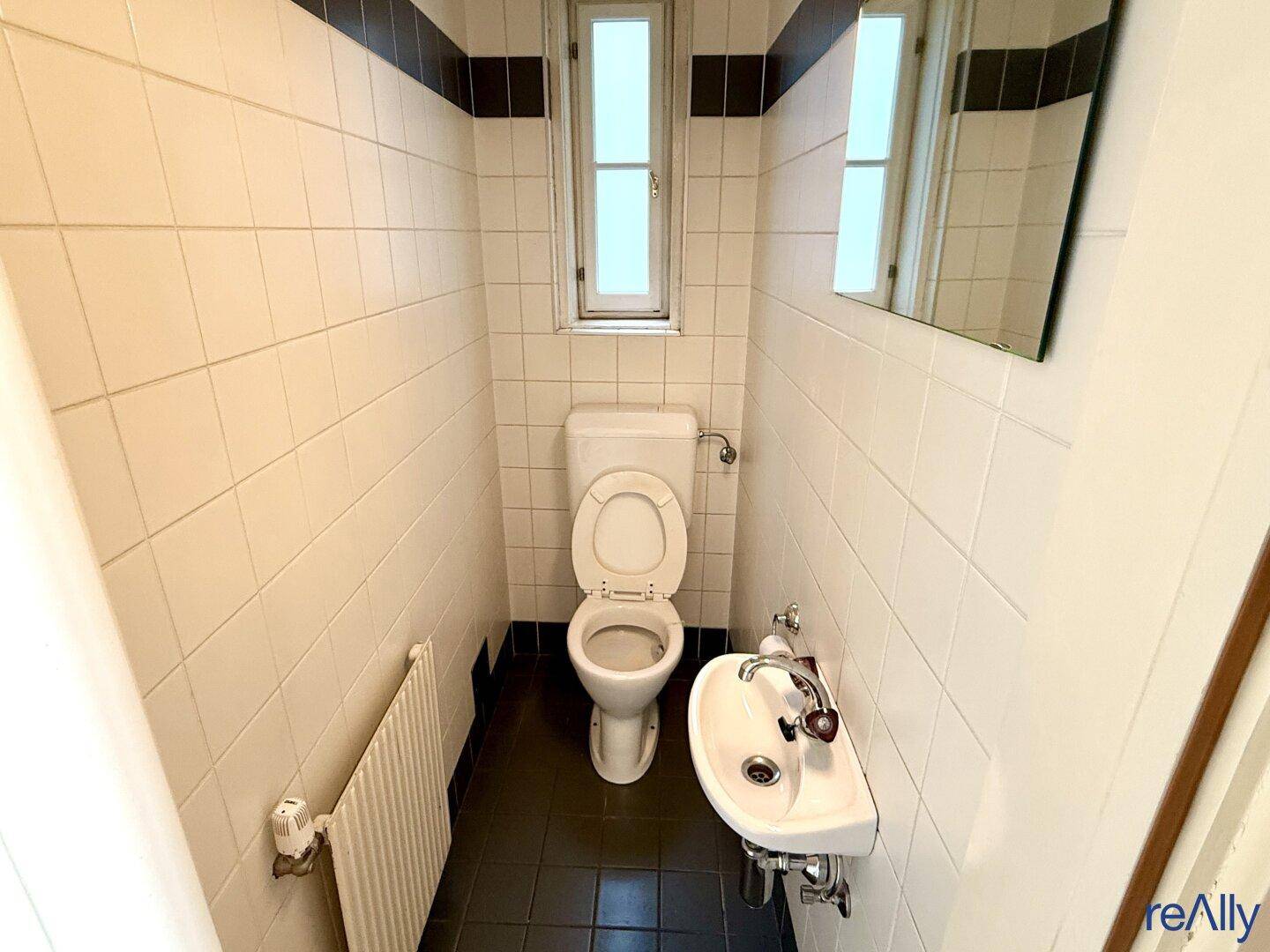 Kleine Gästetoilette mit weißen Fliesen, dunklem Boden und einem Fenster.