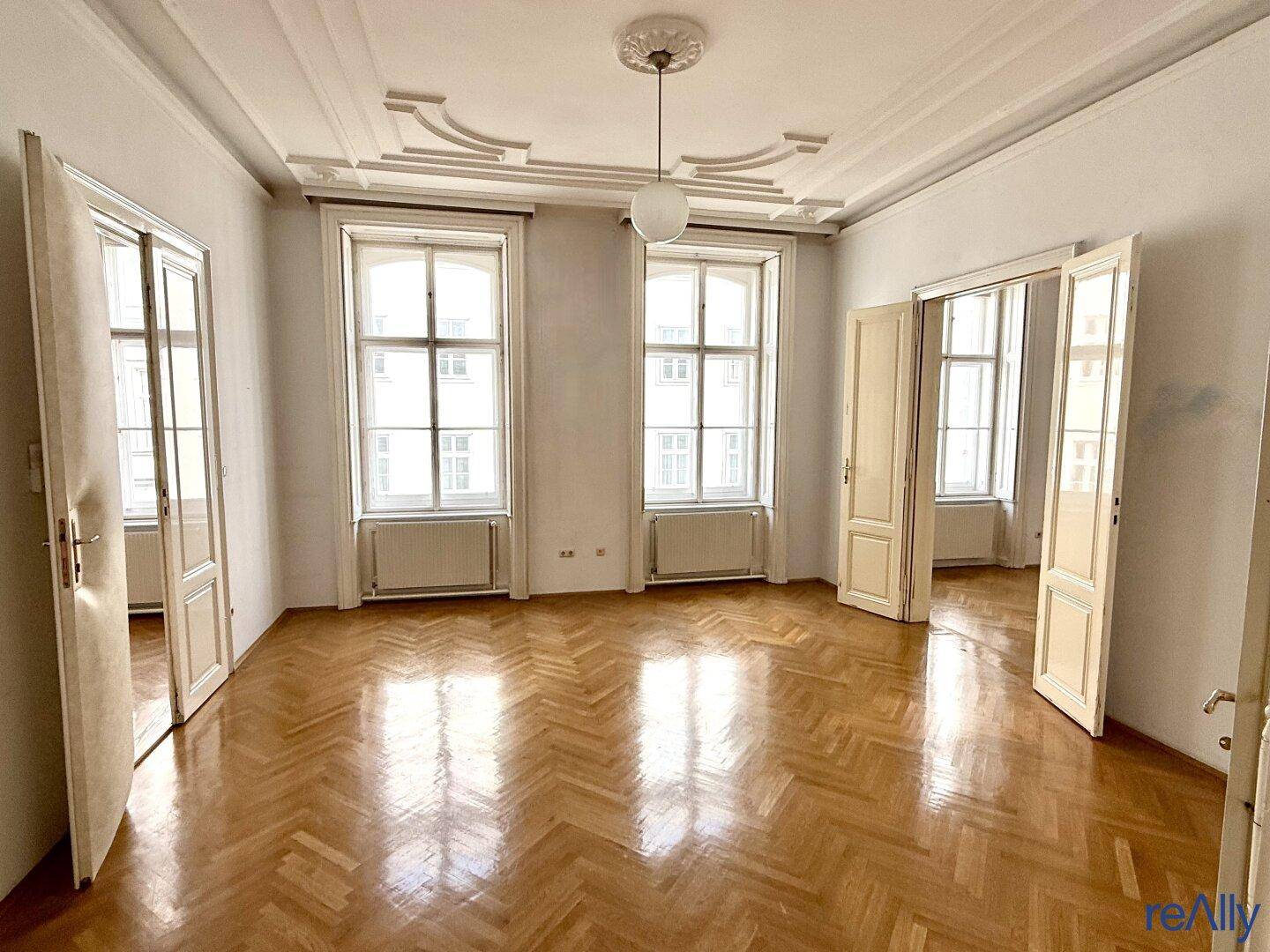 Geräumiges Wohnzimmer mit Fischgrätparkett, hohen Decken und Stuckverzierungen, hell durch große Fenster.