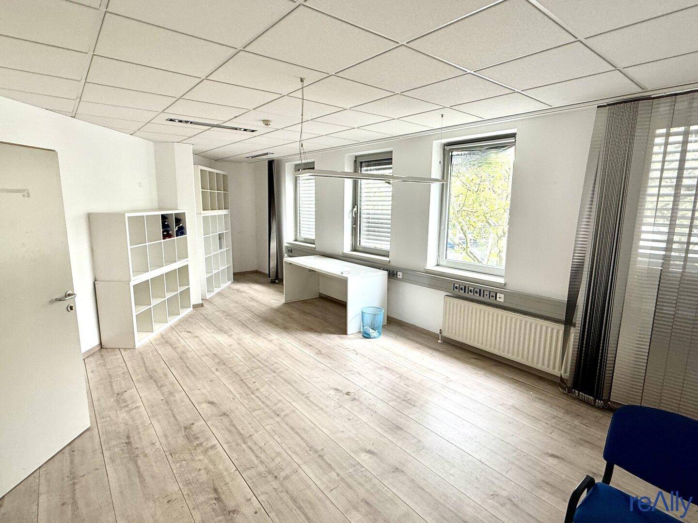 Einzelbüro mit hellem Laminatboden, Fenster mit Jalousien und praktischen Regalen.