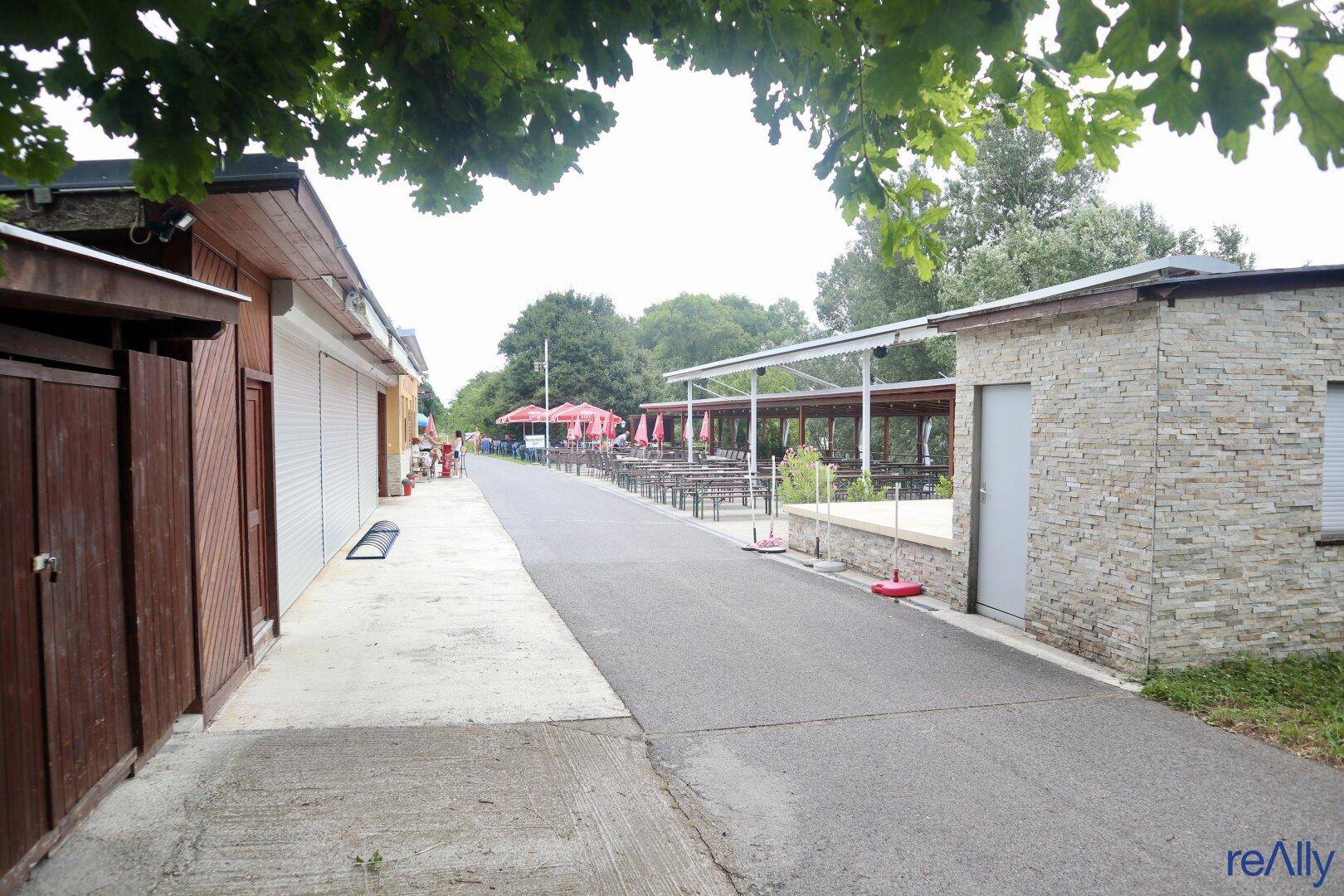 Blick entlang eines Weges mit verschiedenen Gebäuden und dem Außenbereich eines Restaurants im Hintergrund.