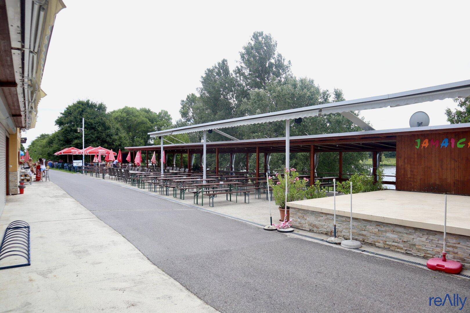 Weitläufiger Außenbereich mit einer Bühne, vielen Biertischen und einer überdachten Terrasse neben einem Gewässer.